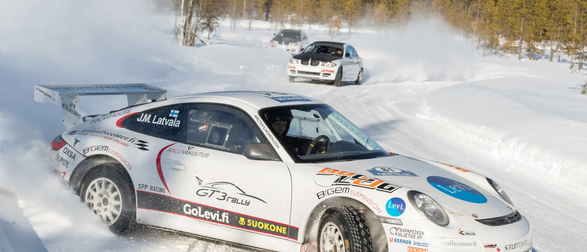 Two rally cars drive fast through a snowy forest track, throwing up clouds of snow behind them.