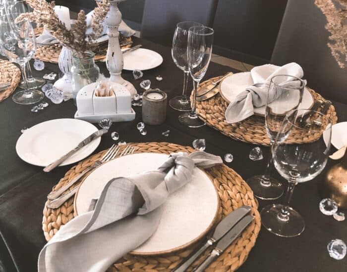 An elegantly set dining table features woven placemats, white plates, folded gray napkins, wine glasses, and scattered decorative crystals.