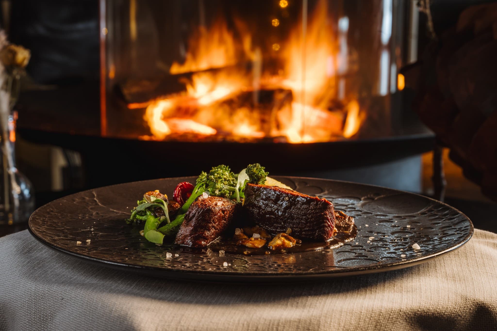 A plated gourmet beef dish is served in front of a glowing fireplace in a dimly lit restaurant setting.