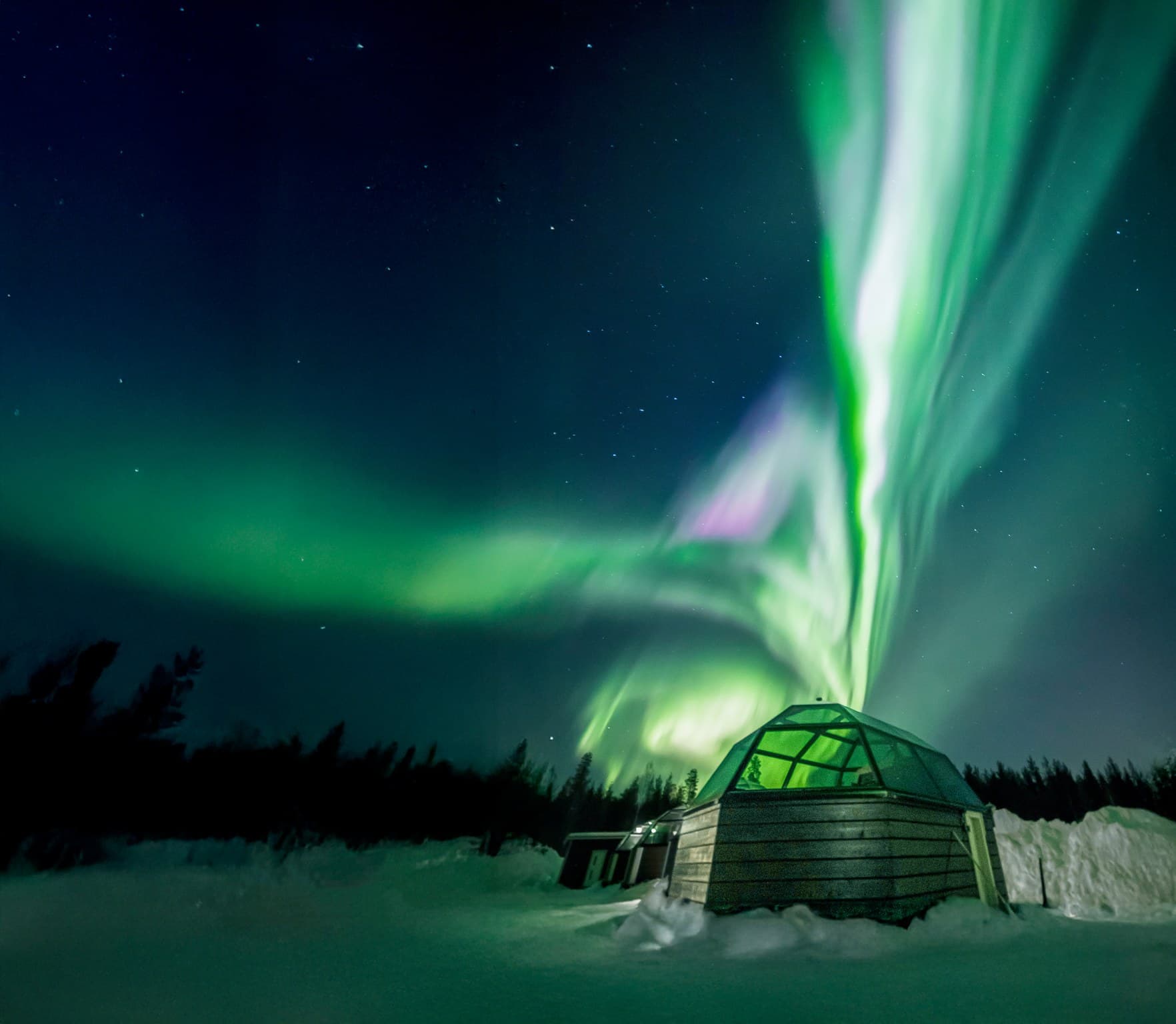Vivid green aurora borealis over a snow-covered landscape with a glass-roof igloo at night.