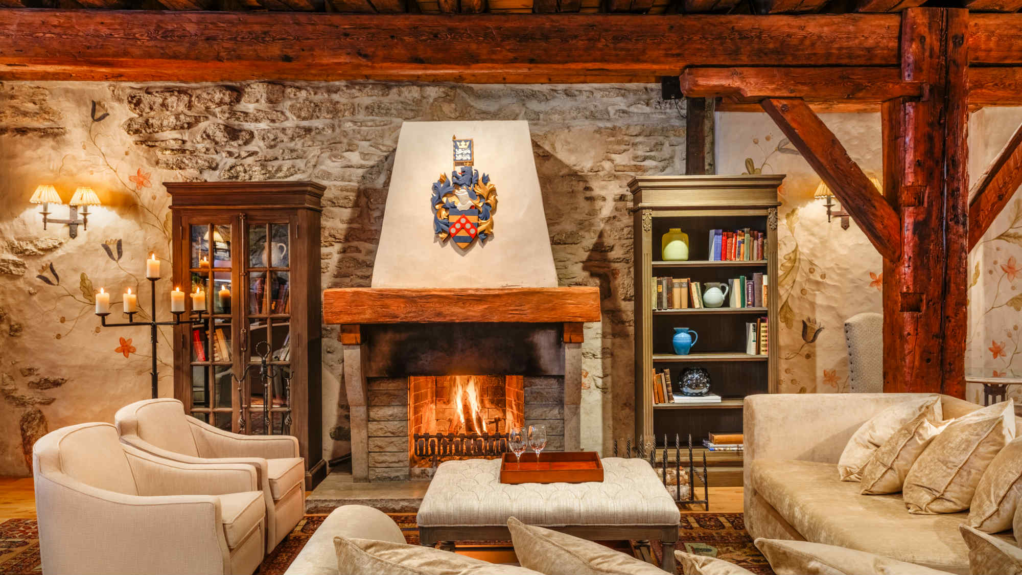 A cozy rustic lounge with a lit stone fireplace, cushioned seating, bookshelves, candles, and exposed wooden beams.