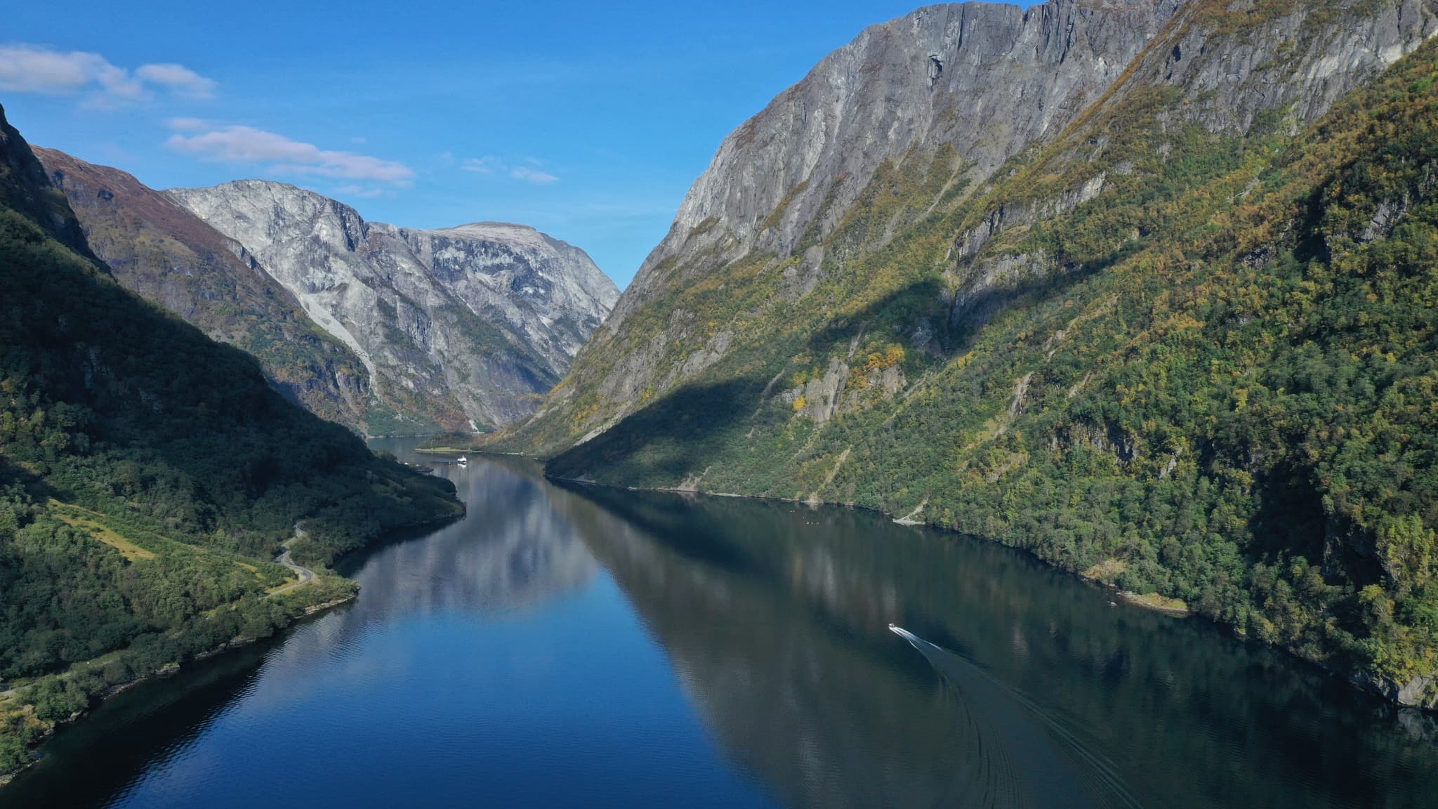 A dramatic fjord landscape with steep rocky mountains, calm dark water, and a small boat leaving a wake beneath a clear blue sky.