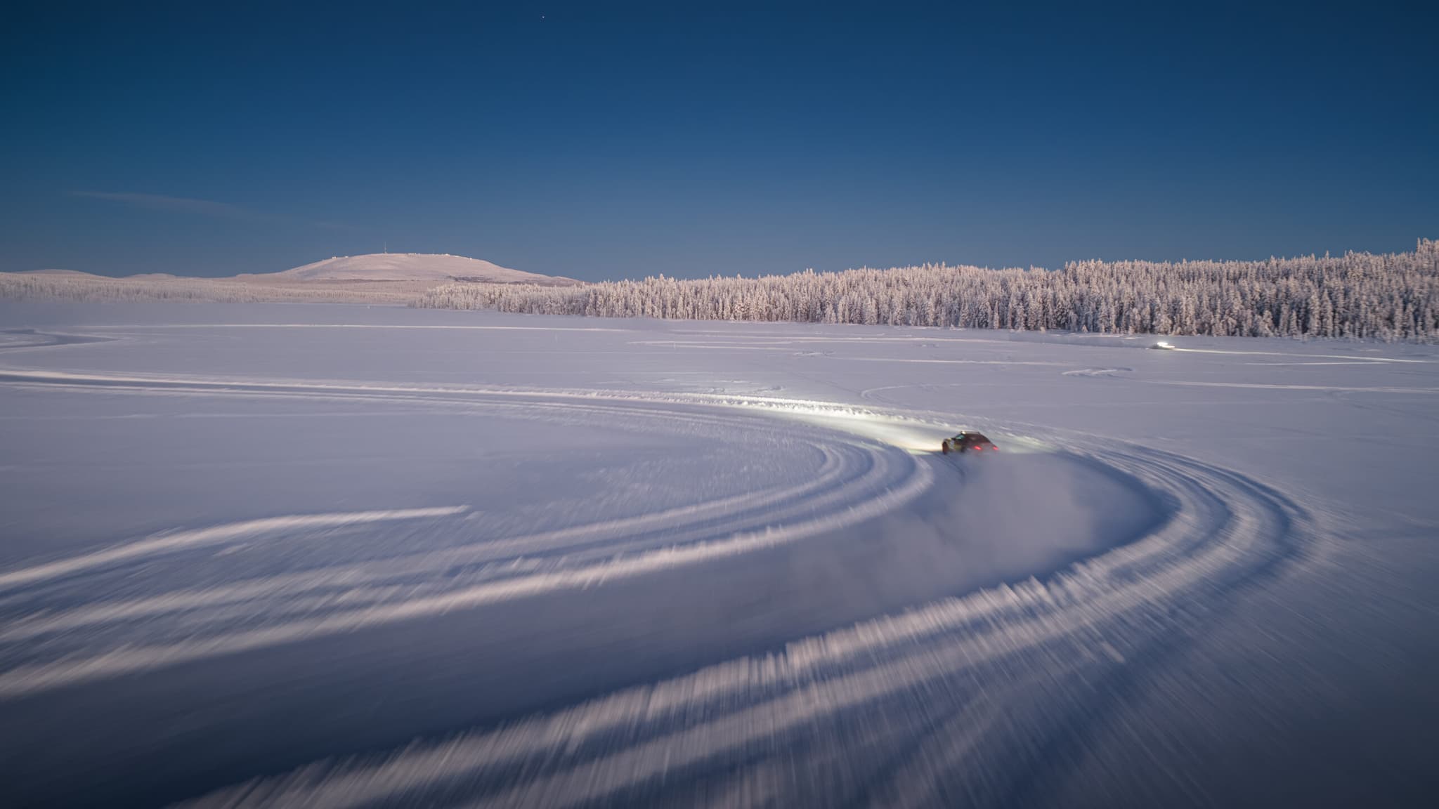A vehicle carving tracks across a snowy frozen landscape beside a frosted forest under a clear blue sky.