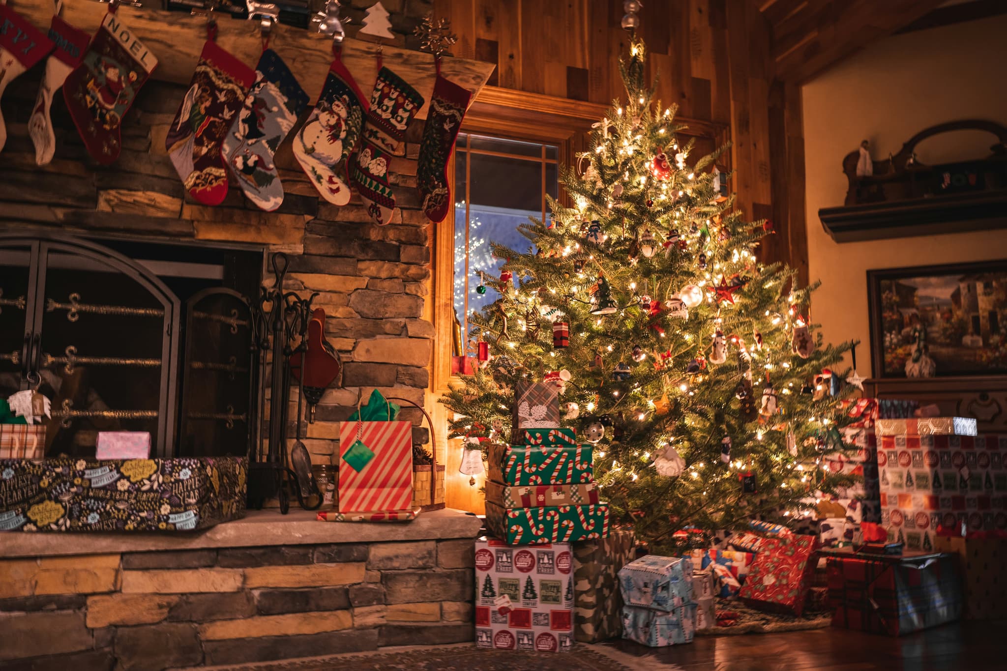A warmly lit living room is decorated for Christmas with a glowing tree, wrapped presents, and stockings hanging above a stone fireplace.