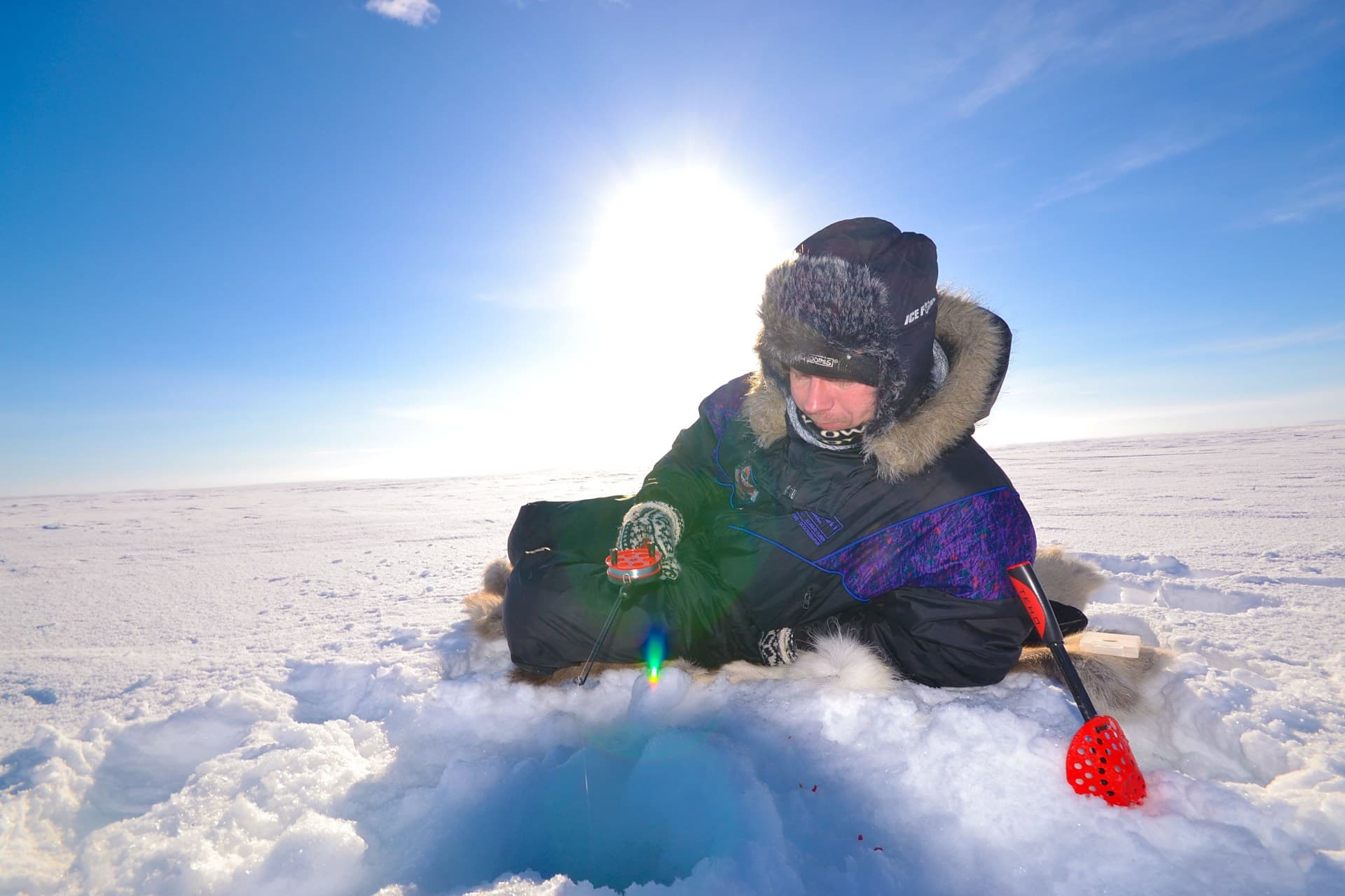 A bundled-up person sits on the snow while ice fishing on a frozen landscape under a bright sun.