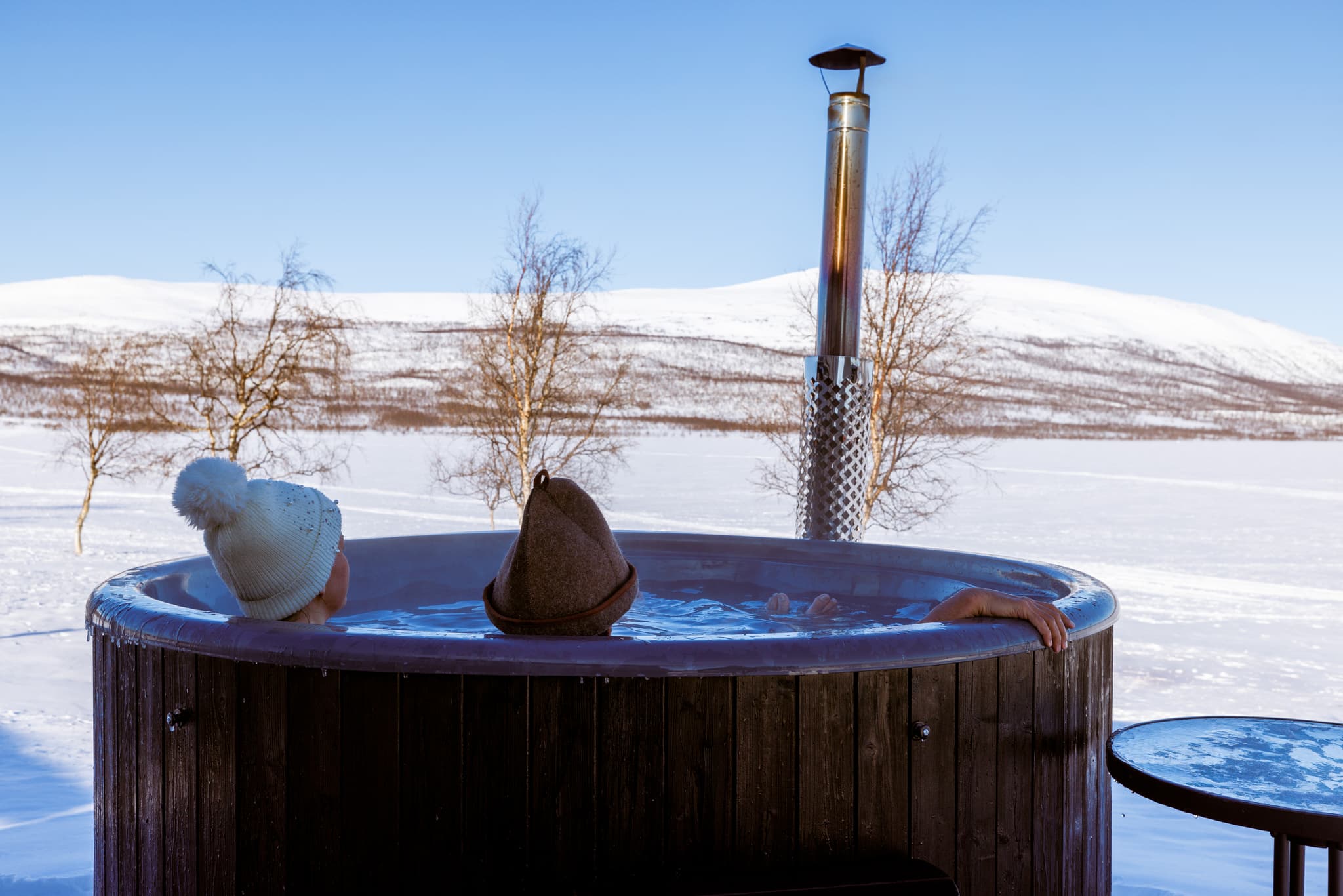 Two people relax in a round outdoor hot tub in a snowy landscape with bare trees and a clear blue sky.