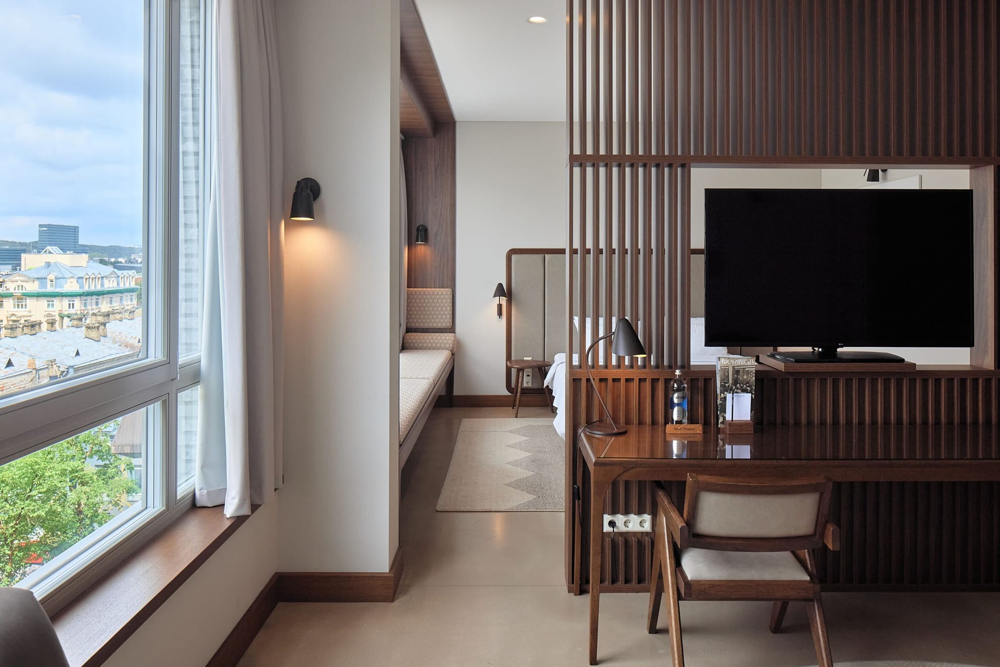 A modern hotel room with a large window view, a wooden desk, a TV, and a partially separated sleeping area.