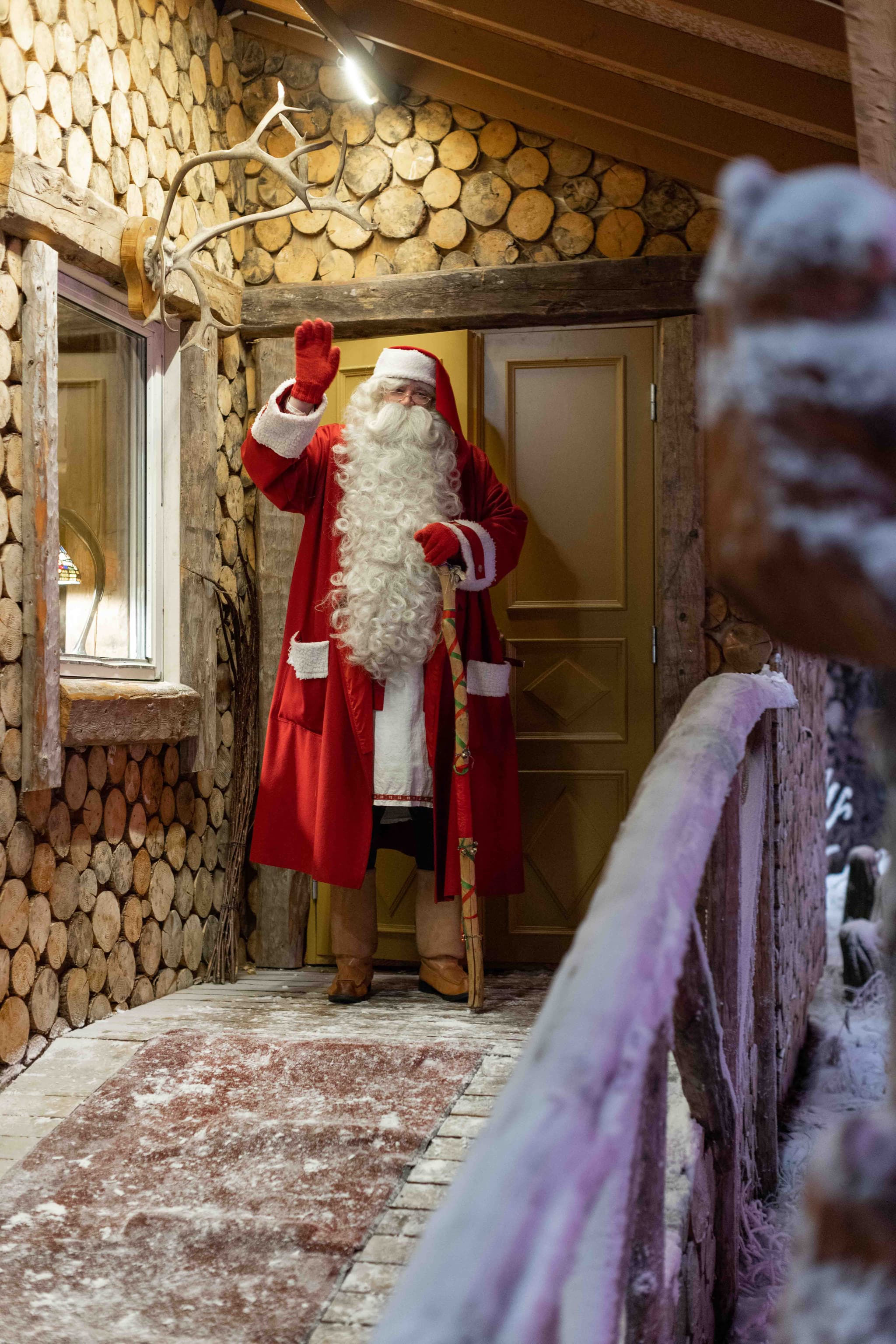 A person dressed as Santa Claus stands in a rustic log-lined doorway and waves in a snowy setting.