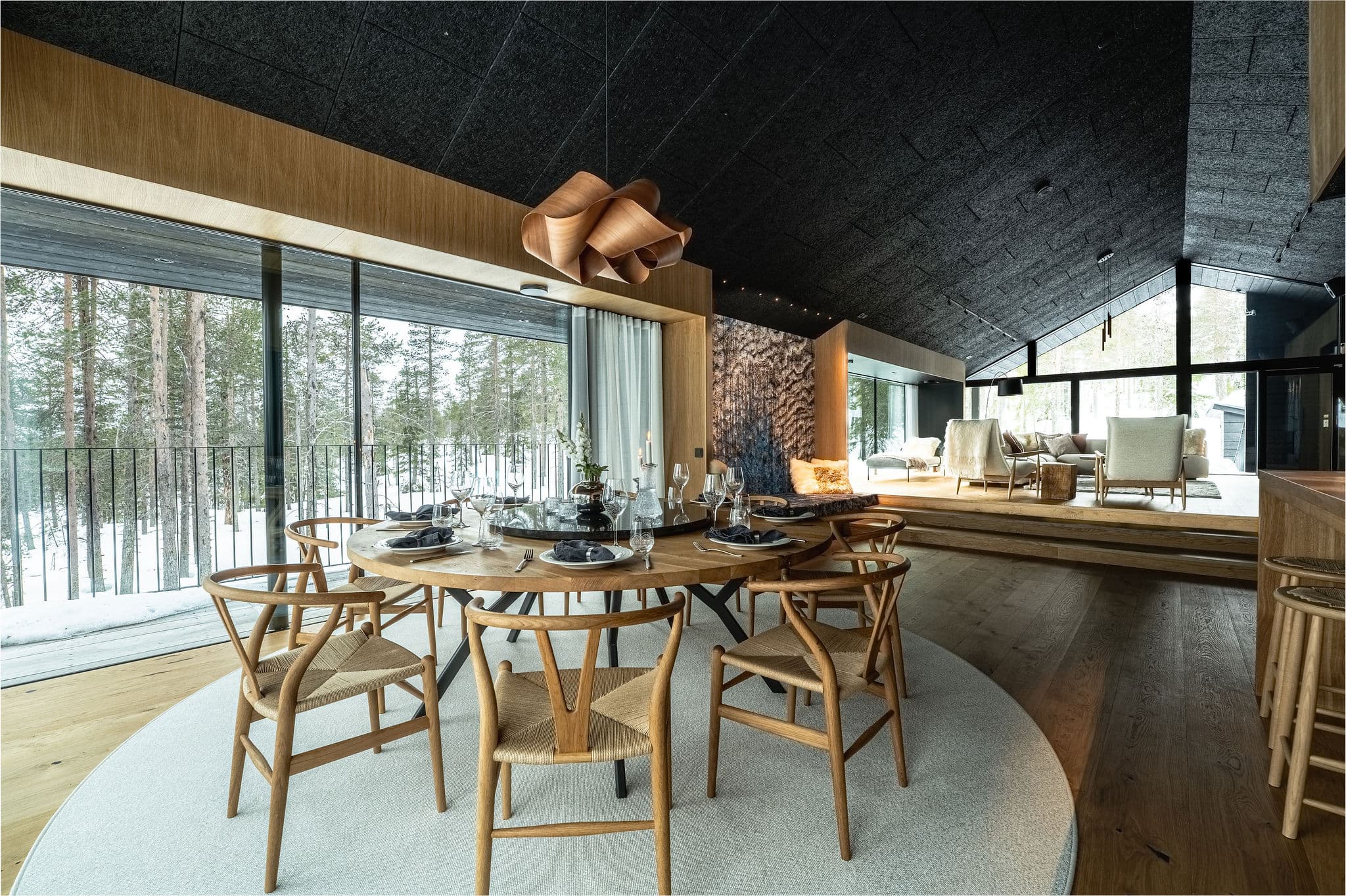 Modern luxury interior with dining table set for a meal, large windows overlooking a snowy forest, and a lounge seating area.