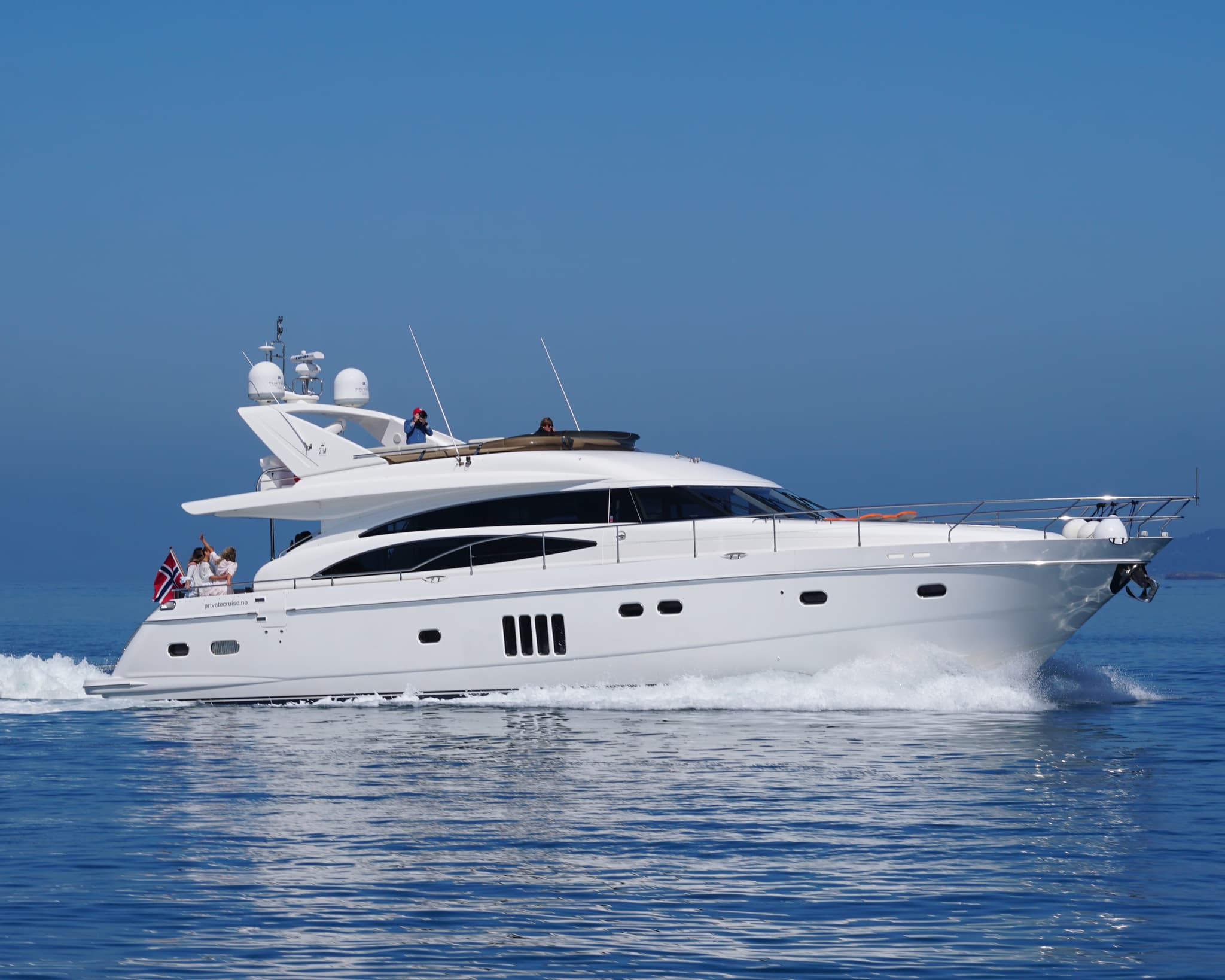 A white motor yacht cruises across calm blue water under a clear sky with a few people visible on deck.