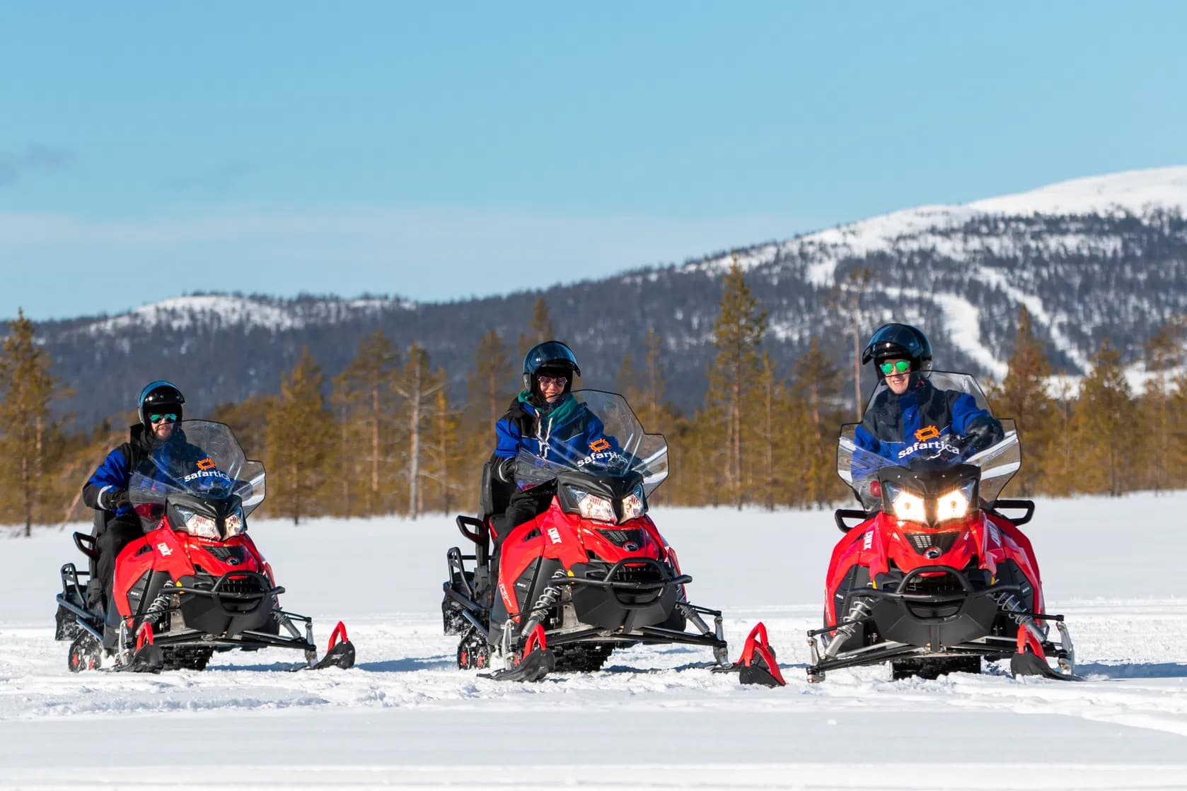 Three people ride red snowmobiles across a snowy landscape with forested mountains in the background.