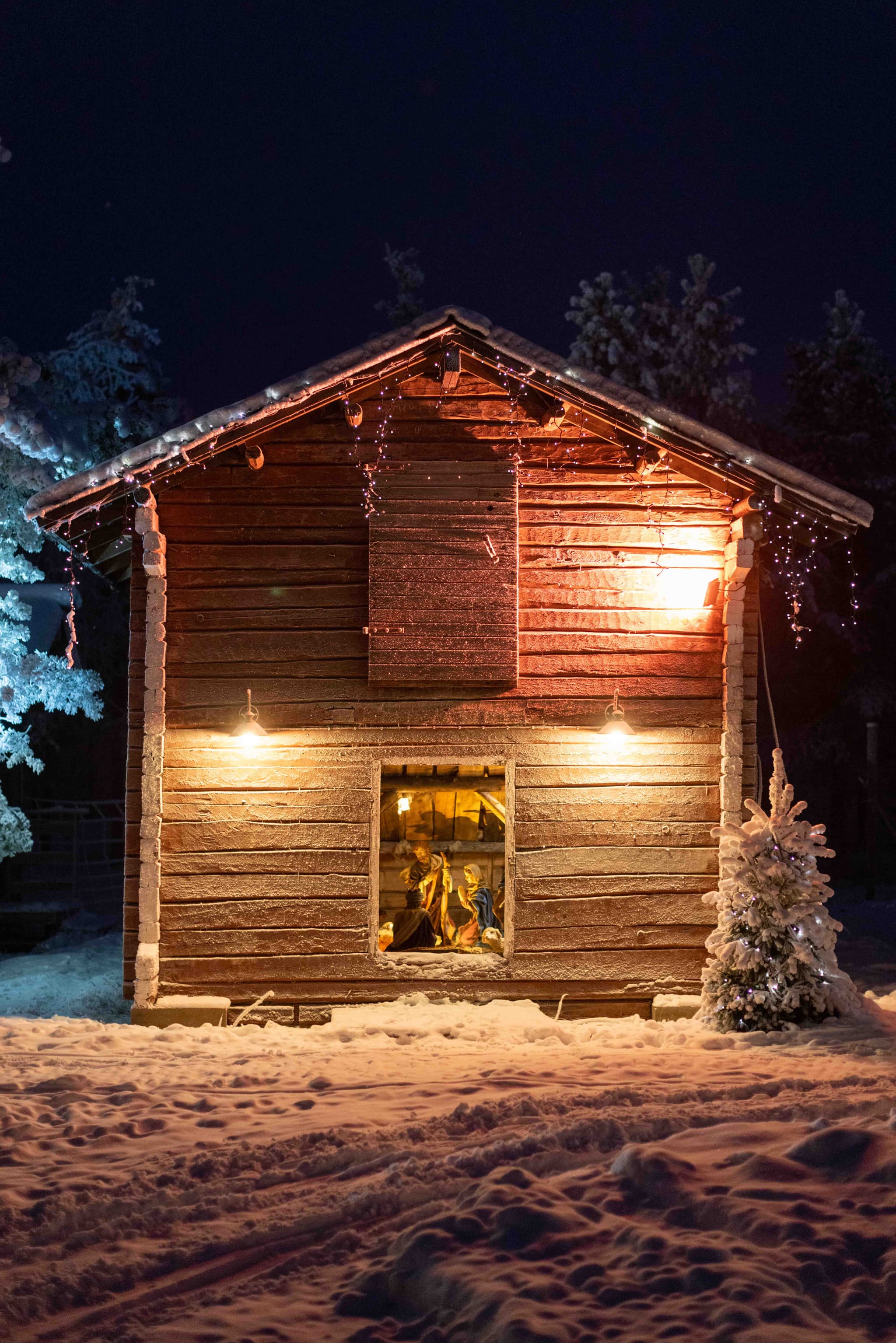 A small wooden cabin decorated with glowing lights stands in a snowy landscape at night, with a lit nativity scene visible in the window.