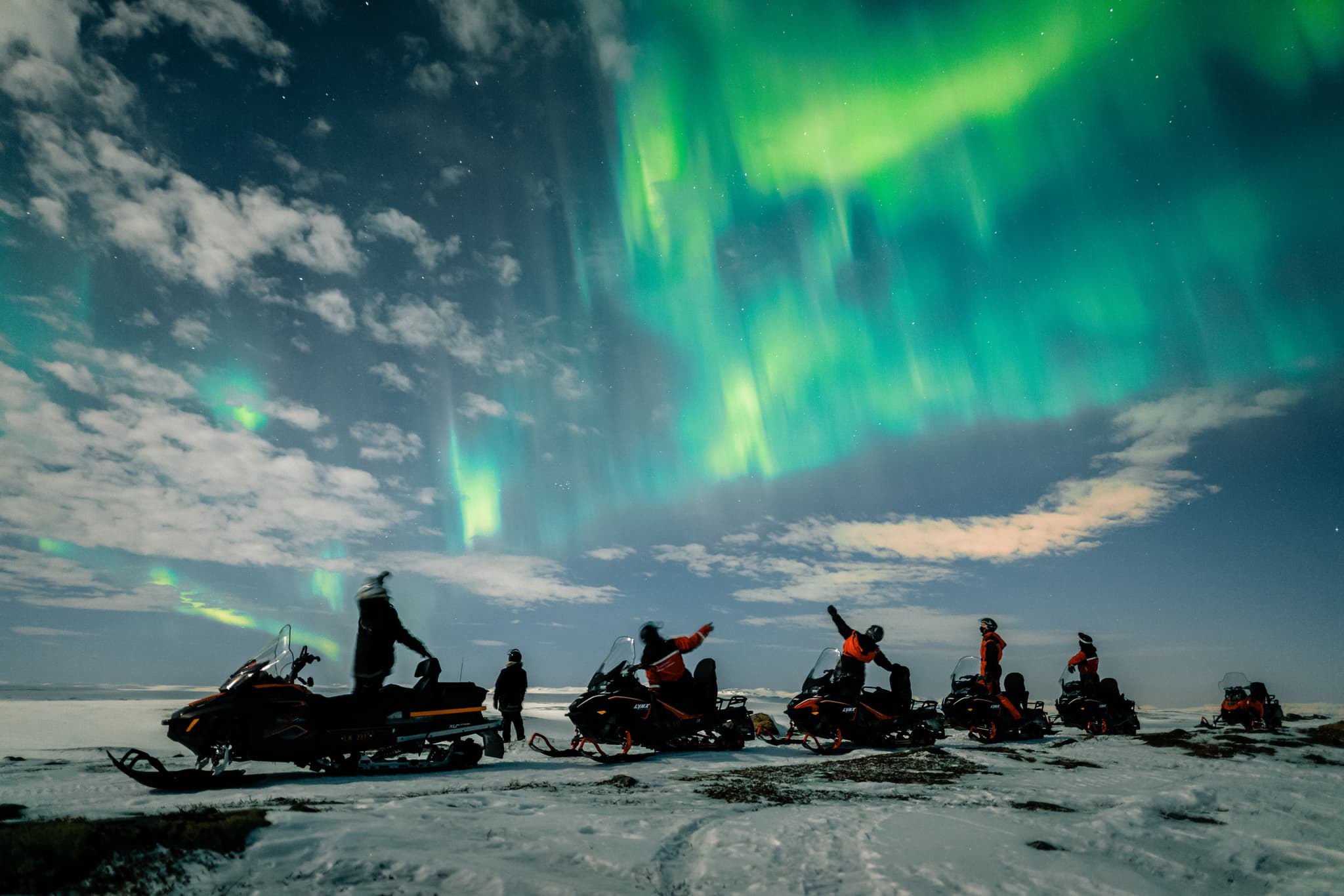 Northern lights over a snow-covered landscape with multiple snowmobiles and riders in winter gear silhouetted in the foreground.