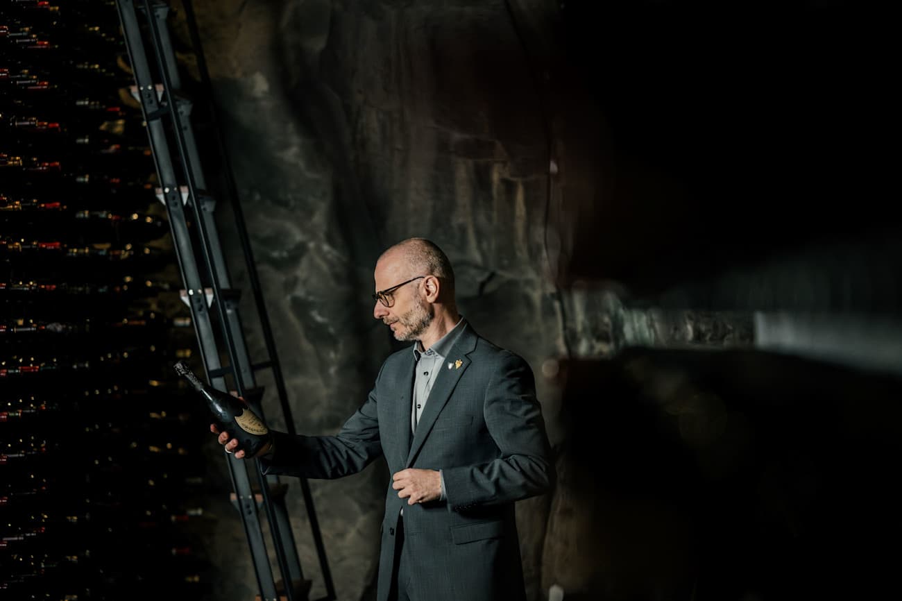 Man in a dark suit holding a champagne bottle while overlooking a nighttime city skyline.