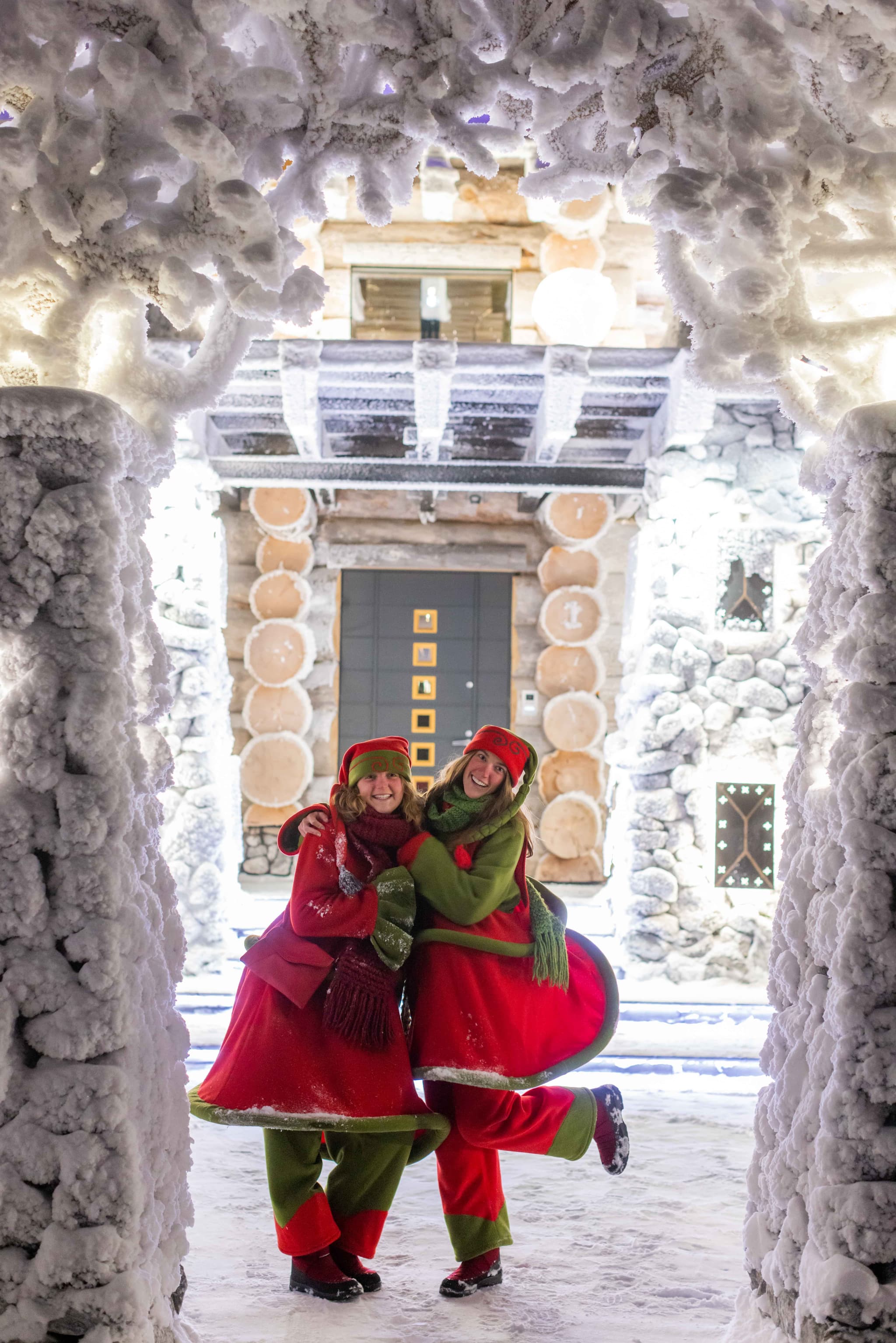 Two people in red and green winter costumes pose smiling in a snow-covered entrance framed by thick frost and an illuminated rustic building behind them.