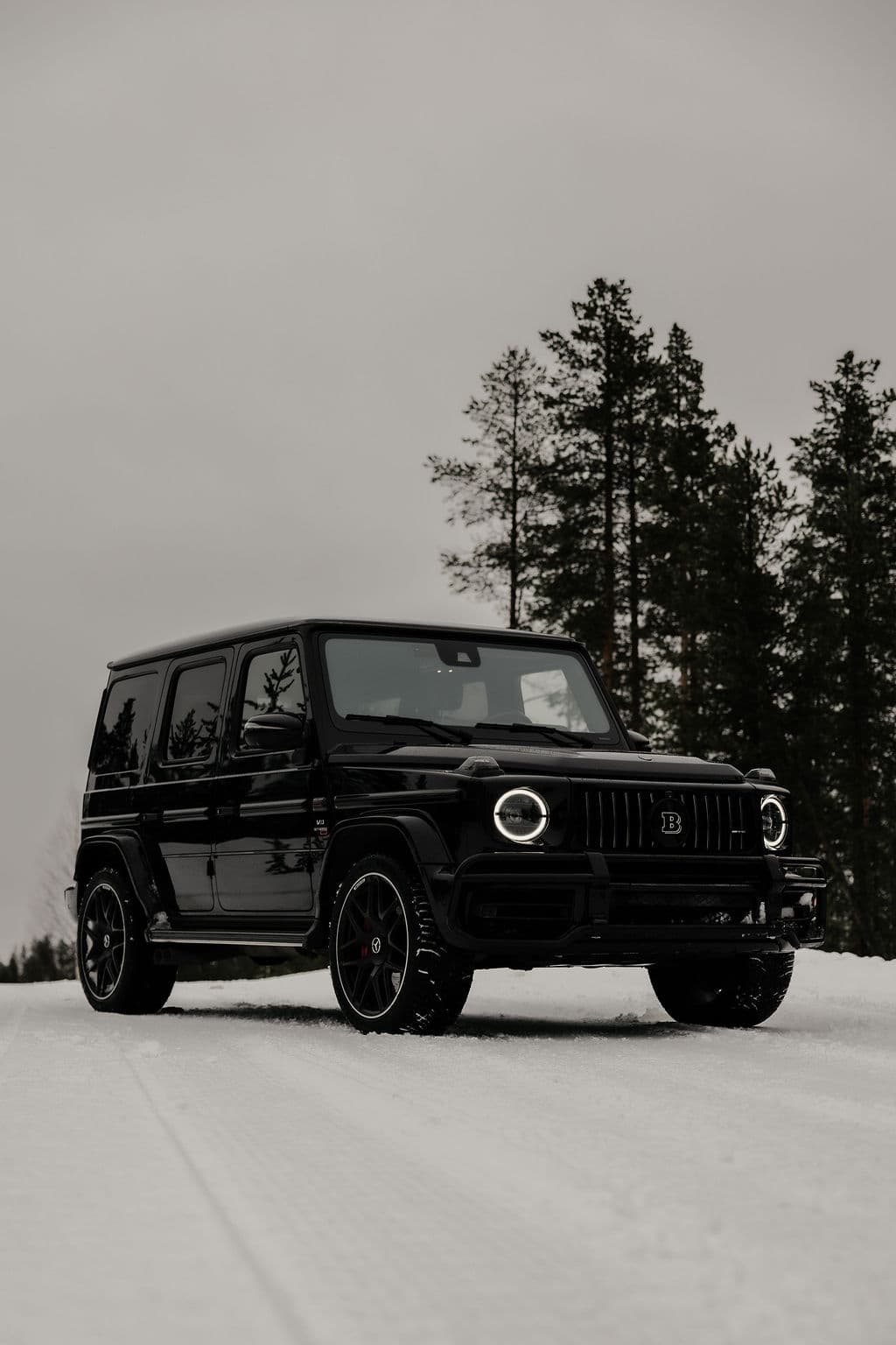 A black luxury SUV is parked on a snowy road with tall evergreen trees in the background under an overcast sky.