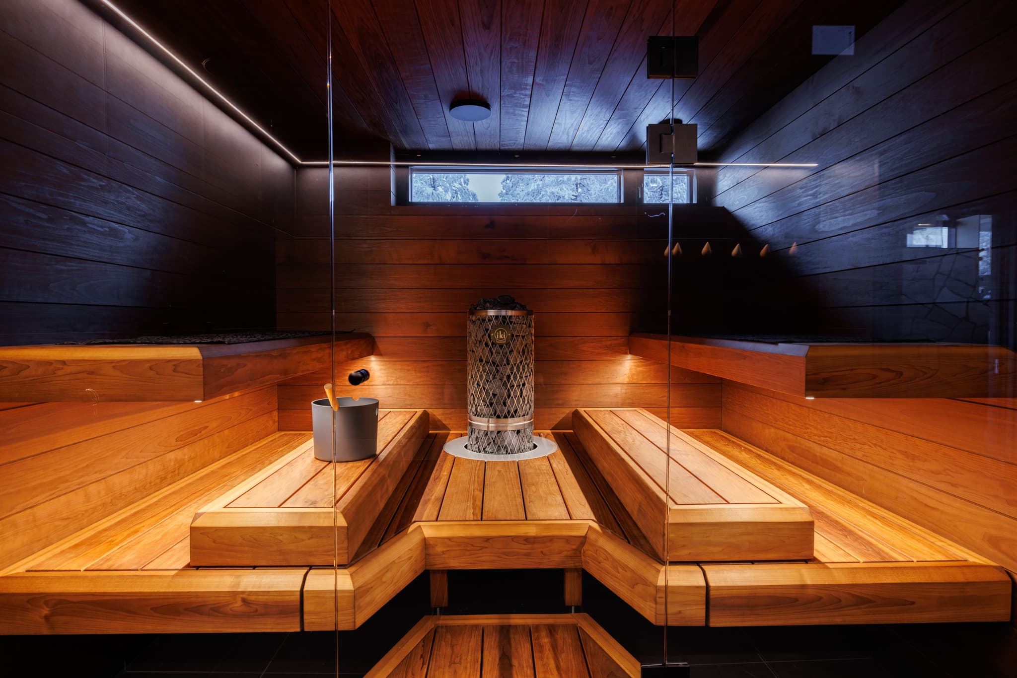 A modern wooden sauna room with glass walls, built-in benches, and a central heater.
