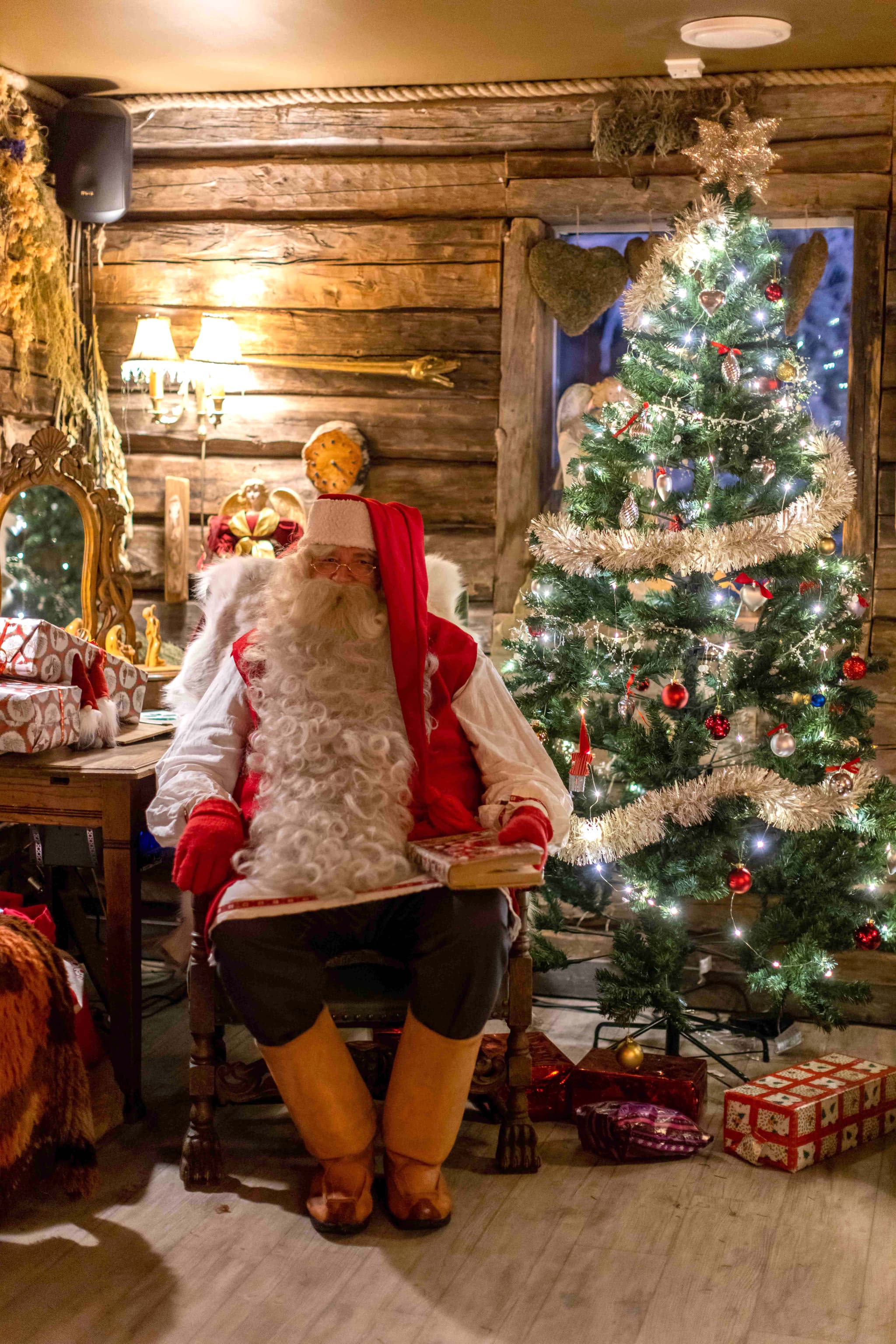 A Santa Claus figure sits in a rustic room beside a lit Christmas tree and wrapped gifts.