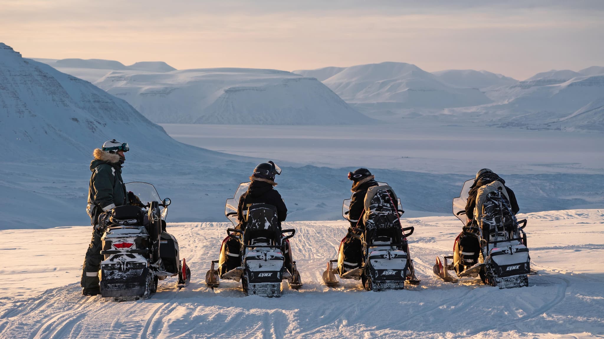 Four people on snowmobiles pause on a snowy mountain overlook with a wide frozen landscape stretching into the distance at sunset.
