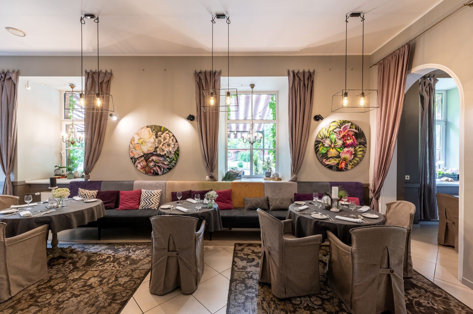 A stylish dining room is set with round tables, upholstered chairs, and floral wall art beneath modern pendant lights.