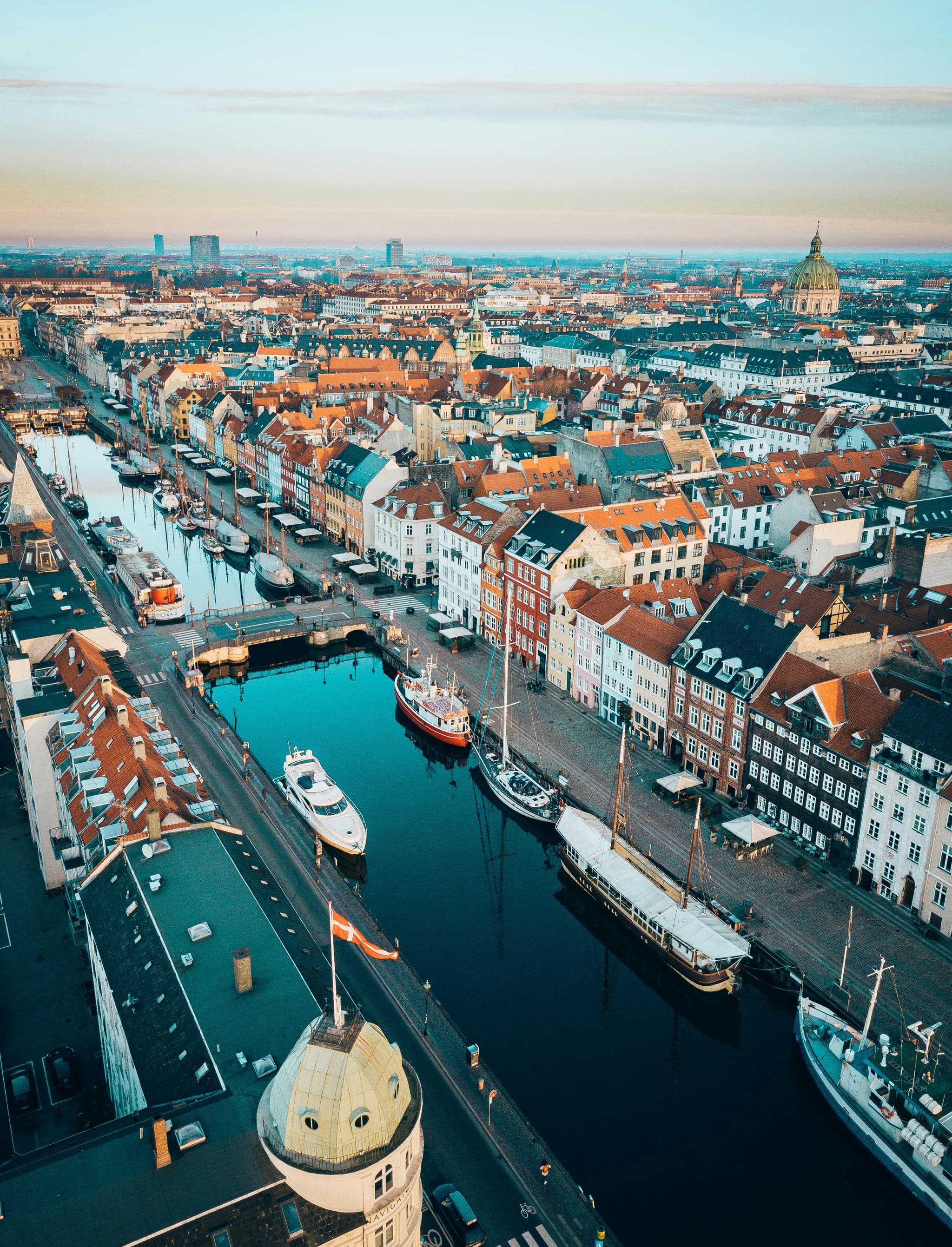 Aerial view of a harbor canal lined with colorful historic buildings and moored boats, with a domed church and city skyline in the distance.