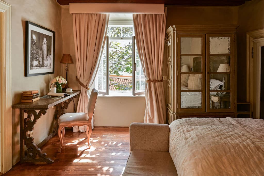 A warmly decorated bedroom with a large window, pink curtains, a wooden desk, and an antique glass cabinet beside the bed.