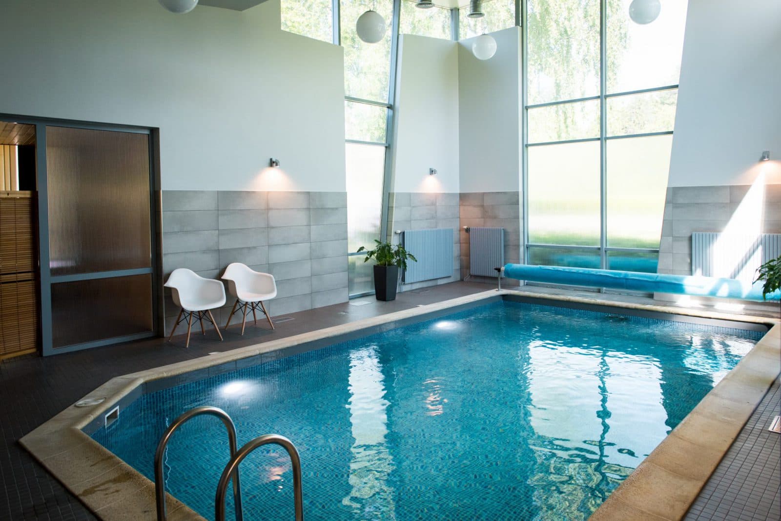 A bright indoor swimming pool with tall windows, blue water, and two white chairs beside the pool deck.