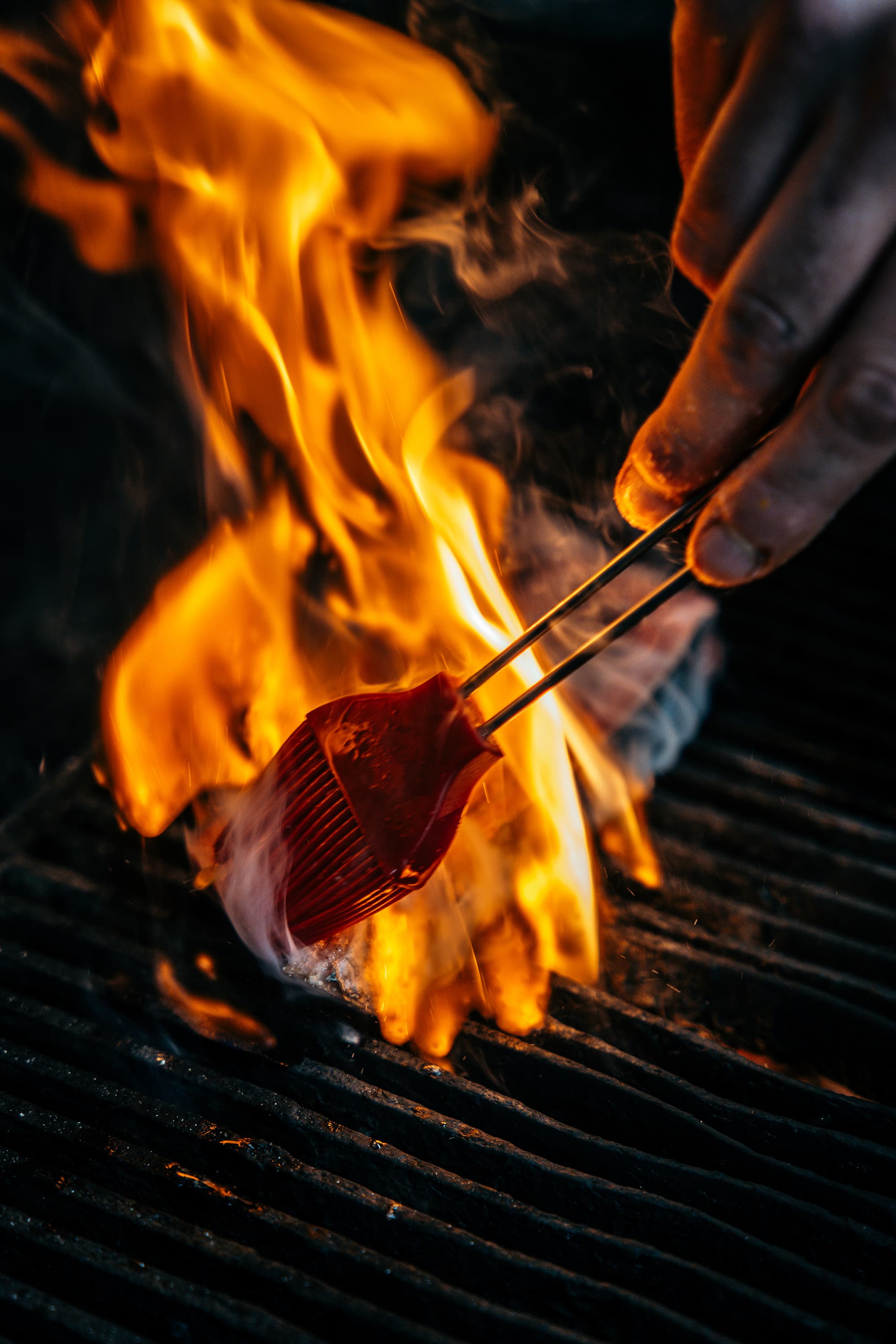 A close-up shows a hand using tongs to hold food over bright flames on a grill.