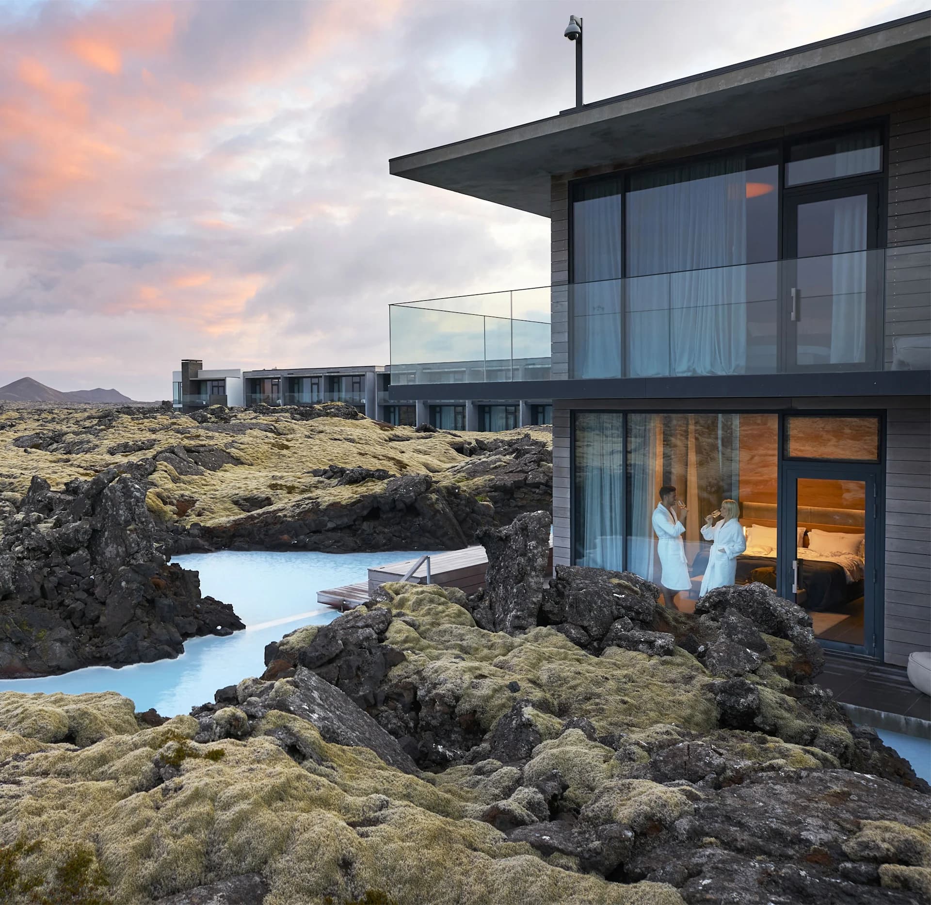 A modern hotel suite overlooks a rocky lava landscape and a pale blue outdoor pool at sunset, with two people in robes standing by the window inside.