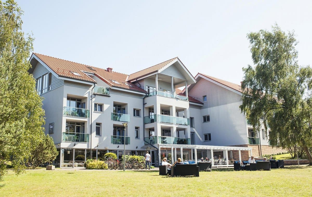 A modern multi-story hotel building with balconies is shown on a sunny day, with guests seated on an outdoor lawn terrace in front.