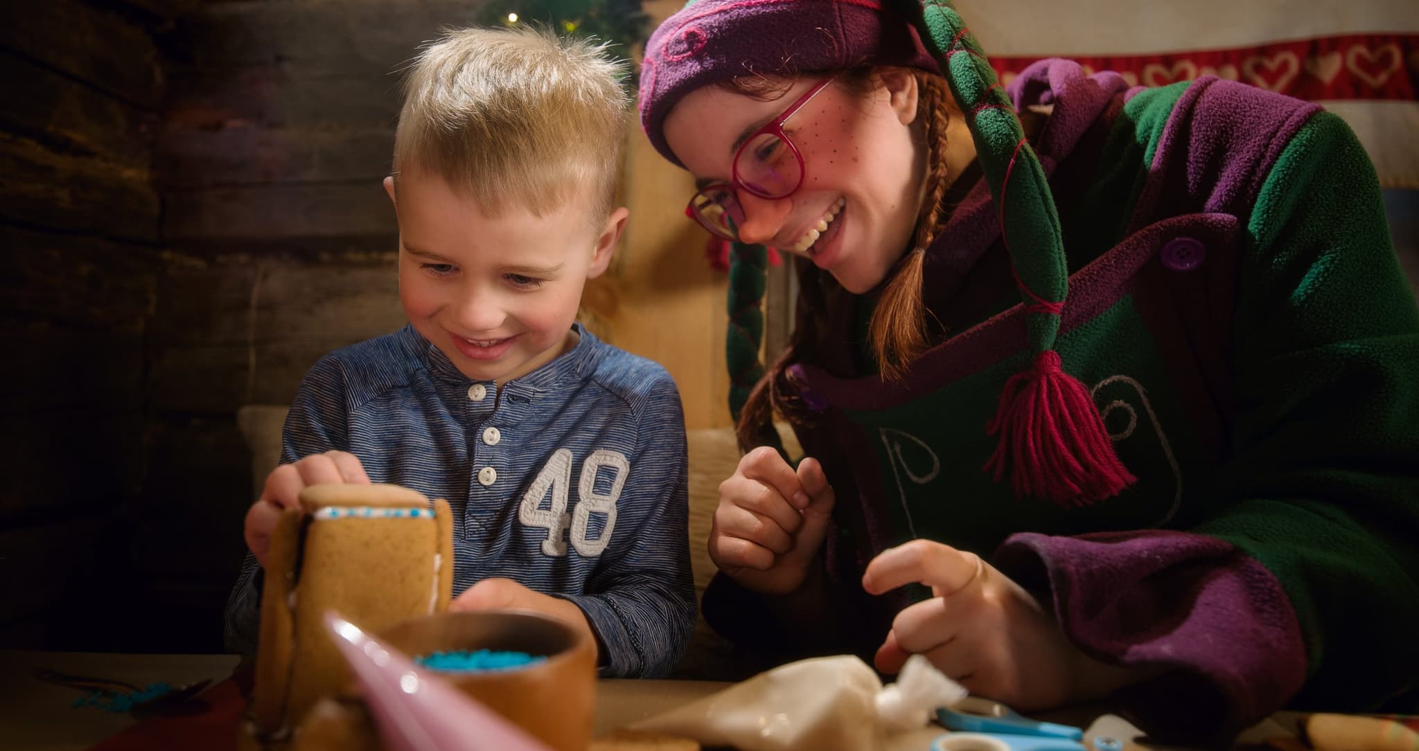 A smiling child and an adult decorate a gingerbread house together at a festive indoor table.