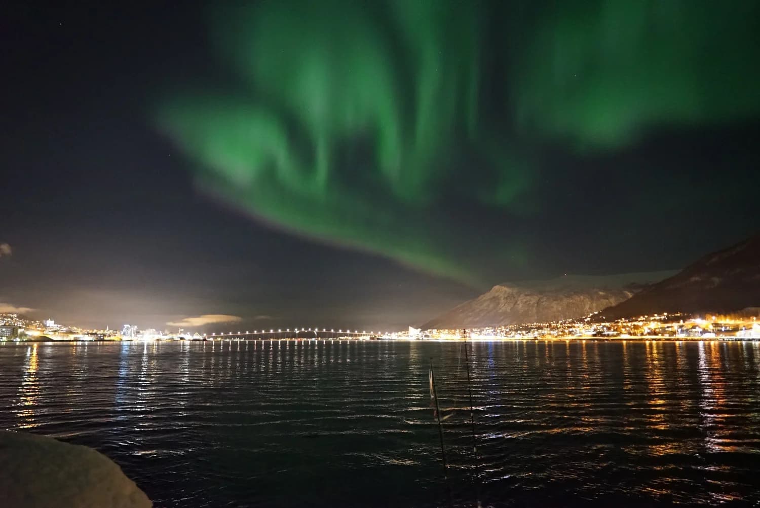 Green northern lights glow across a night sky above a lit coastal city and calm water.