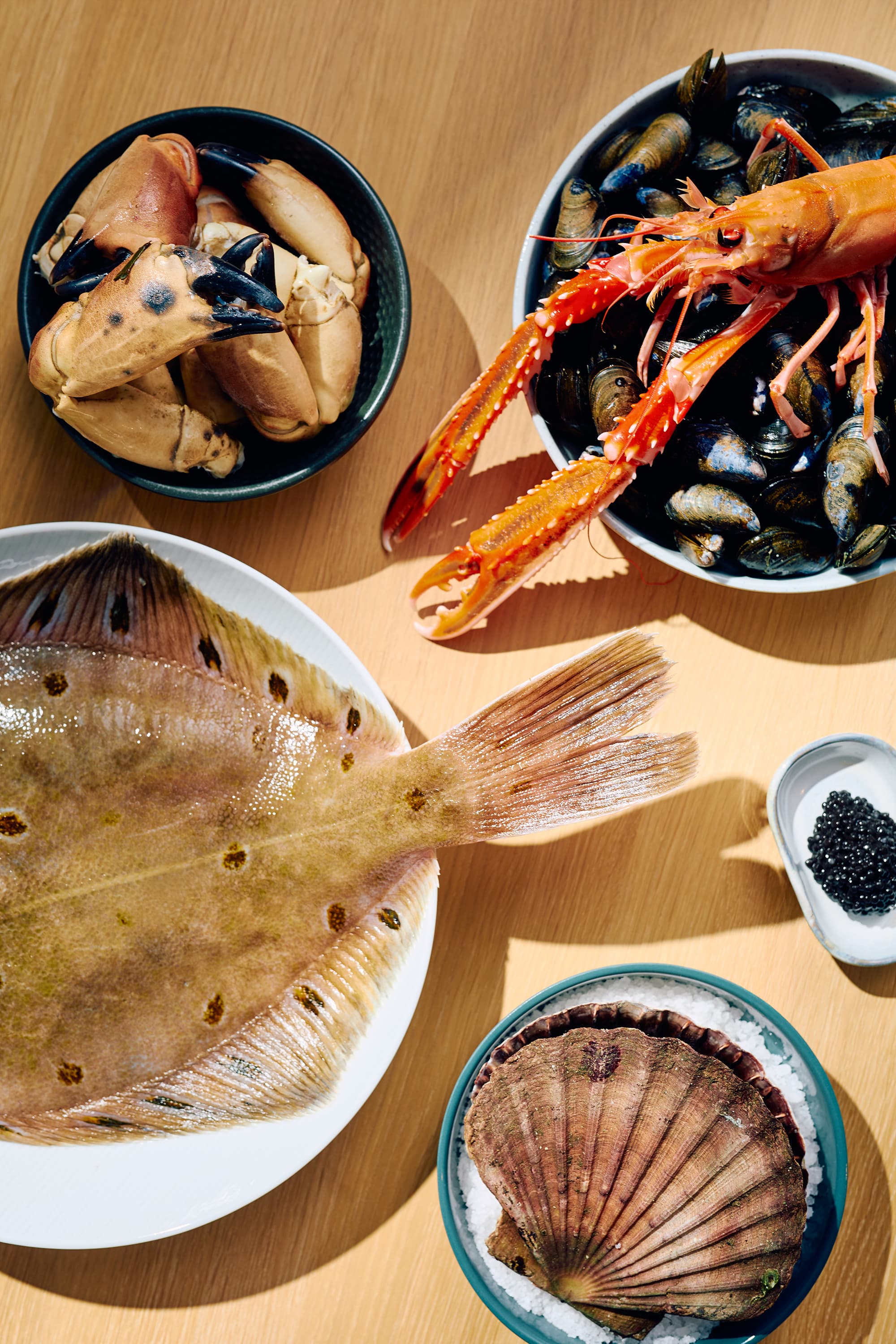An overhead view shows an arrangement of raw seafood on a wooden table, including crab claws, langoustine, mussels, caviar, and large shellfish and fish on plates and bowls.