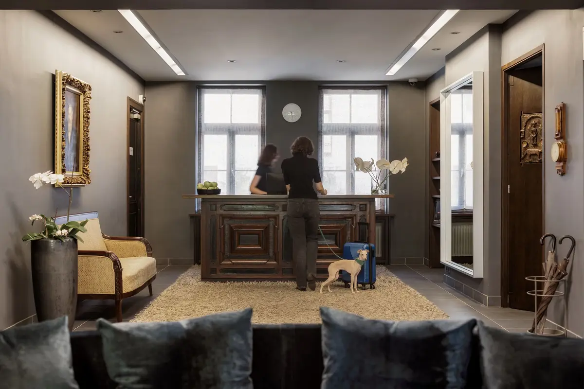 A stylish hotel reception area with two people standing at the front desk, a small dog, and elegant decor.