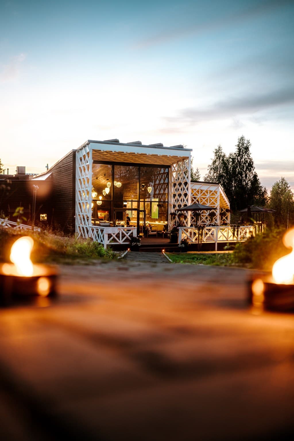 A modern restaurant building with a glass entrance and white lattice trim glows warmly at dusk, framed by soft flames in the foreground.
