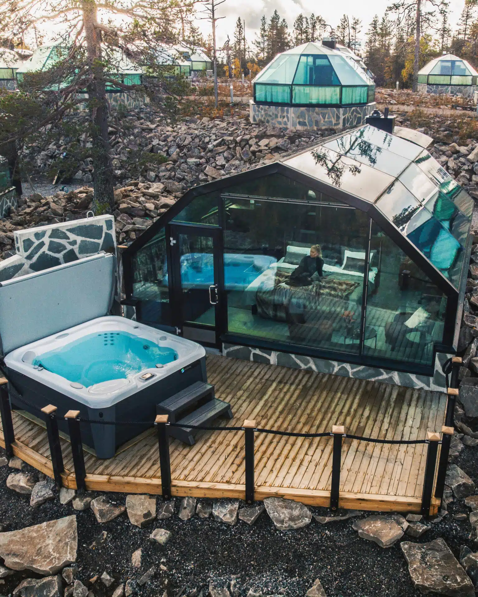 A glass igloo cabin with a private hot tub on a wooden deck is set among rocky ground and trees, with several similar domed cabins in the background.