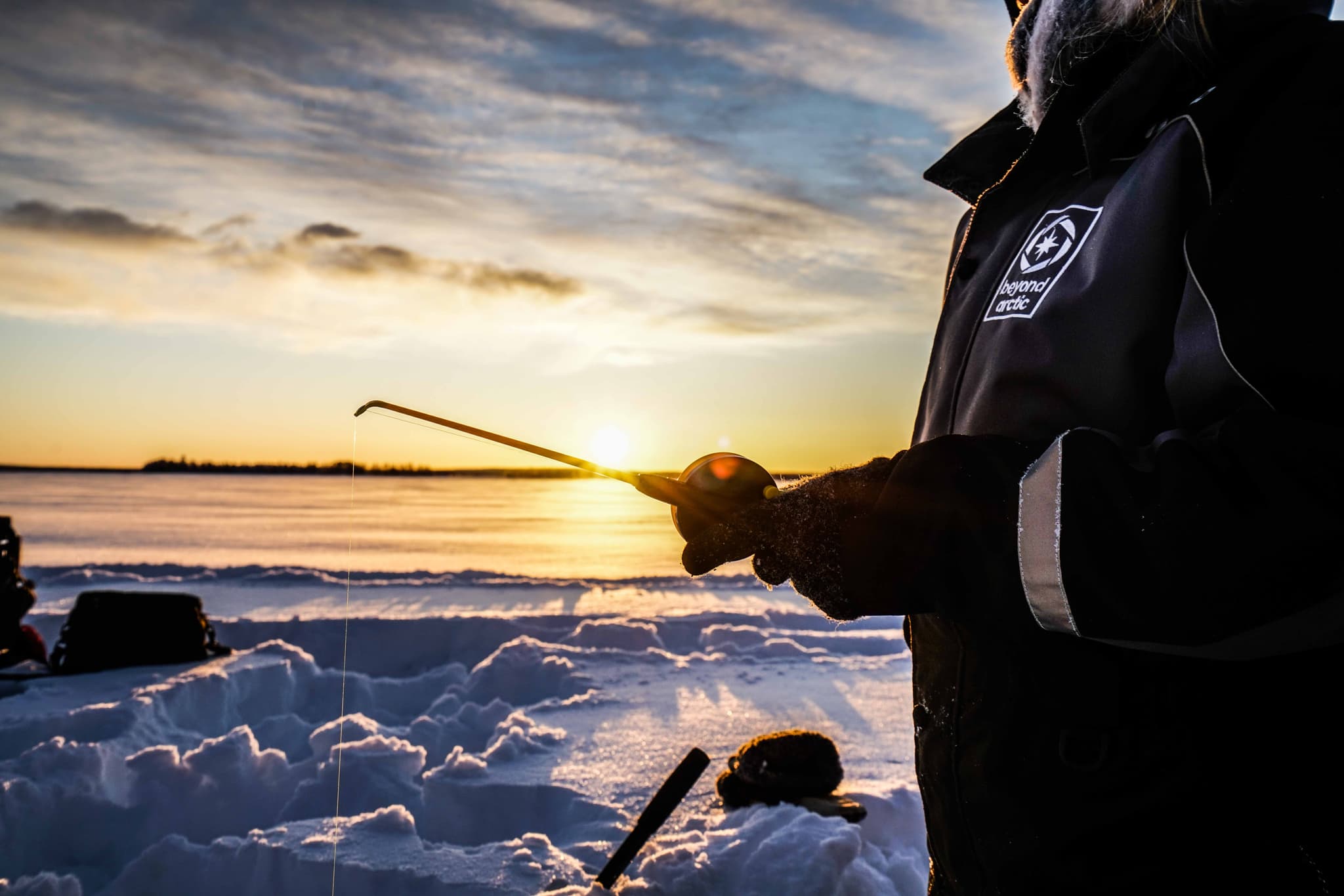 A person in winter clothing holds an ice fishing rod over a hole in the snow beside a frozen lake at sunset.