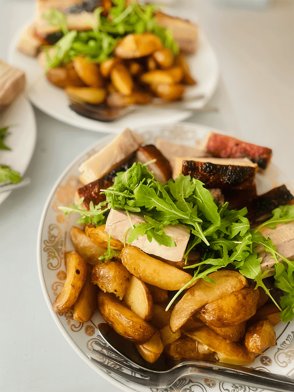 A plate of roasted potato wedges topped with sliced meat and fresh arugula sits on a decorative dish, with another plate blurred in the background.