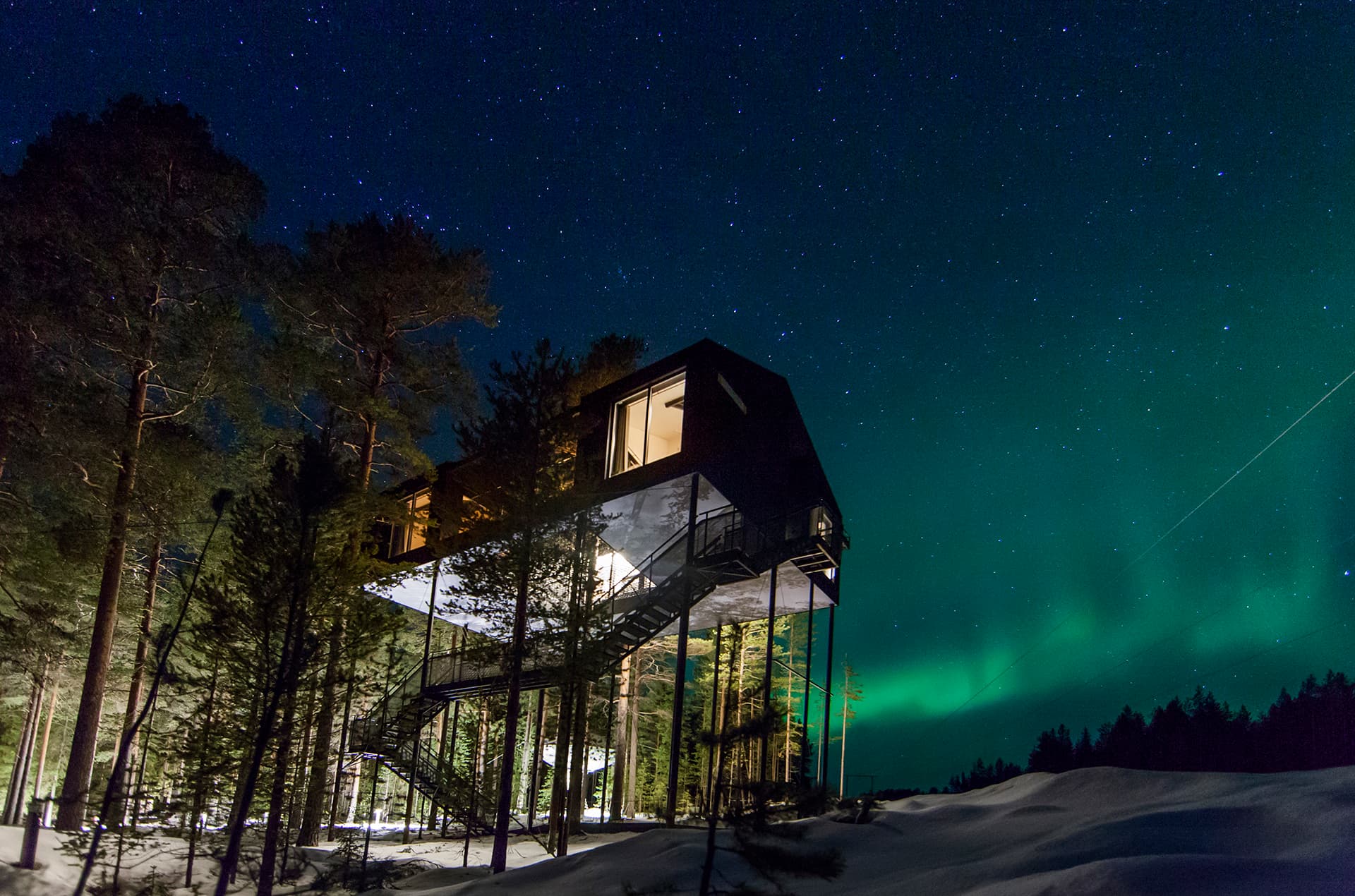 A modern elevated cabin stands among snow-covered trees under a starry night sky with green aurora glowing on the horizon.
