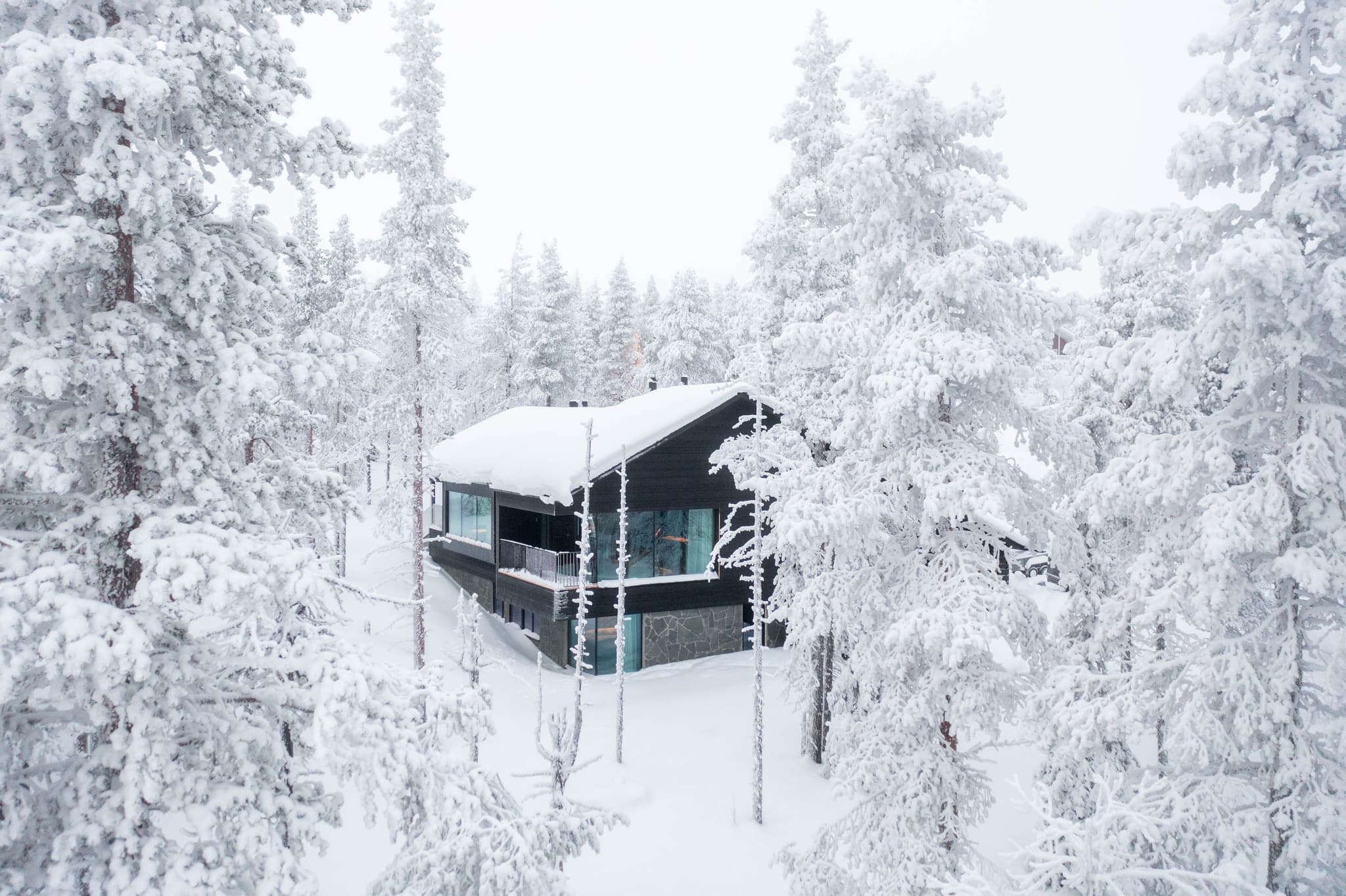 Snow-covered modern cabin nestled in a winter forest, viewed from above with heavy snowfall and frosted trees.
