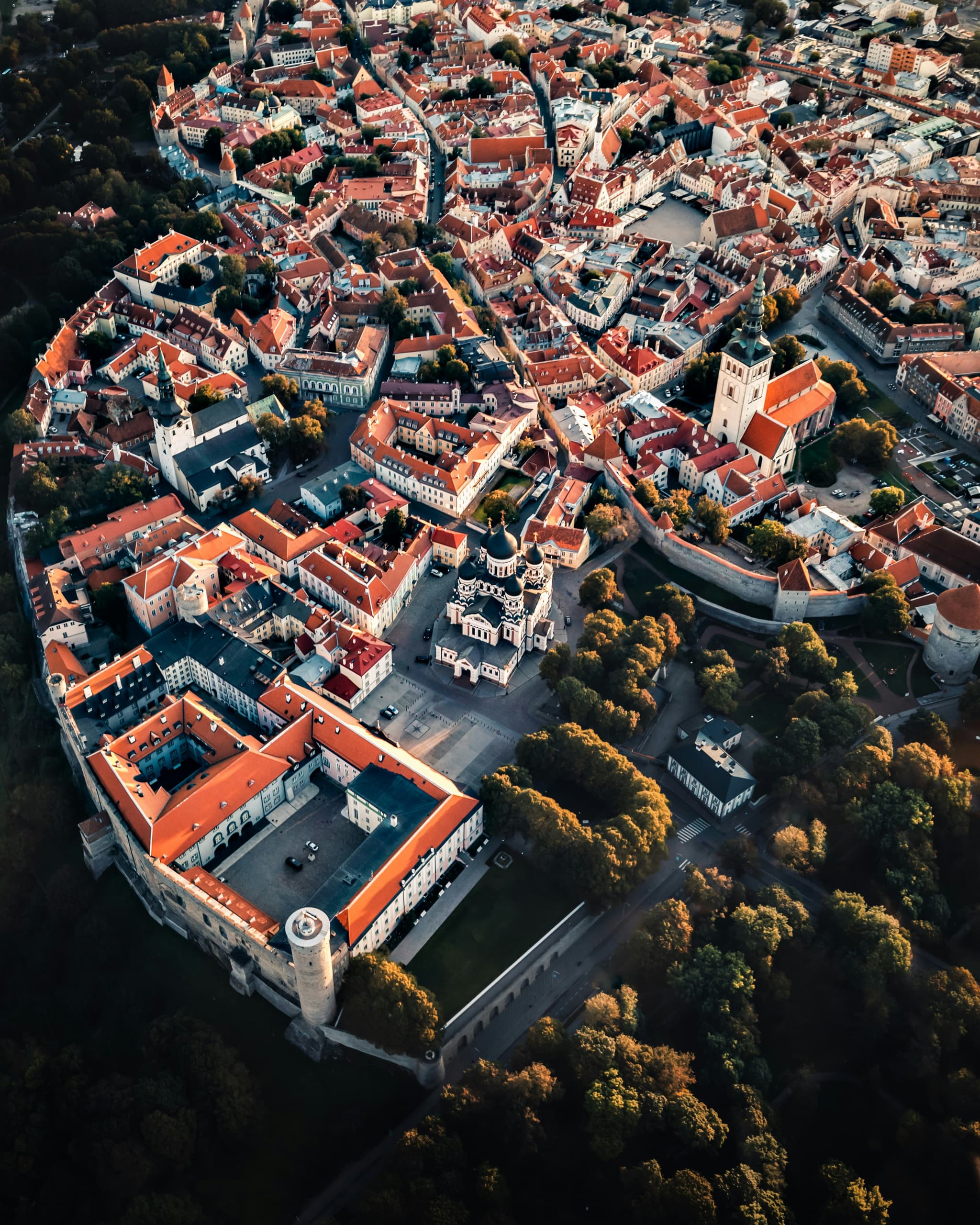 An aerial view shows a historic European old town with dense red-tiled rooftops, church towers, and a surrounding stone wall beside dark green parkland.
