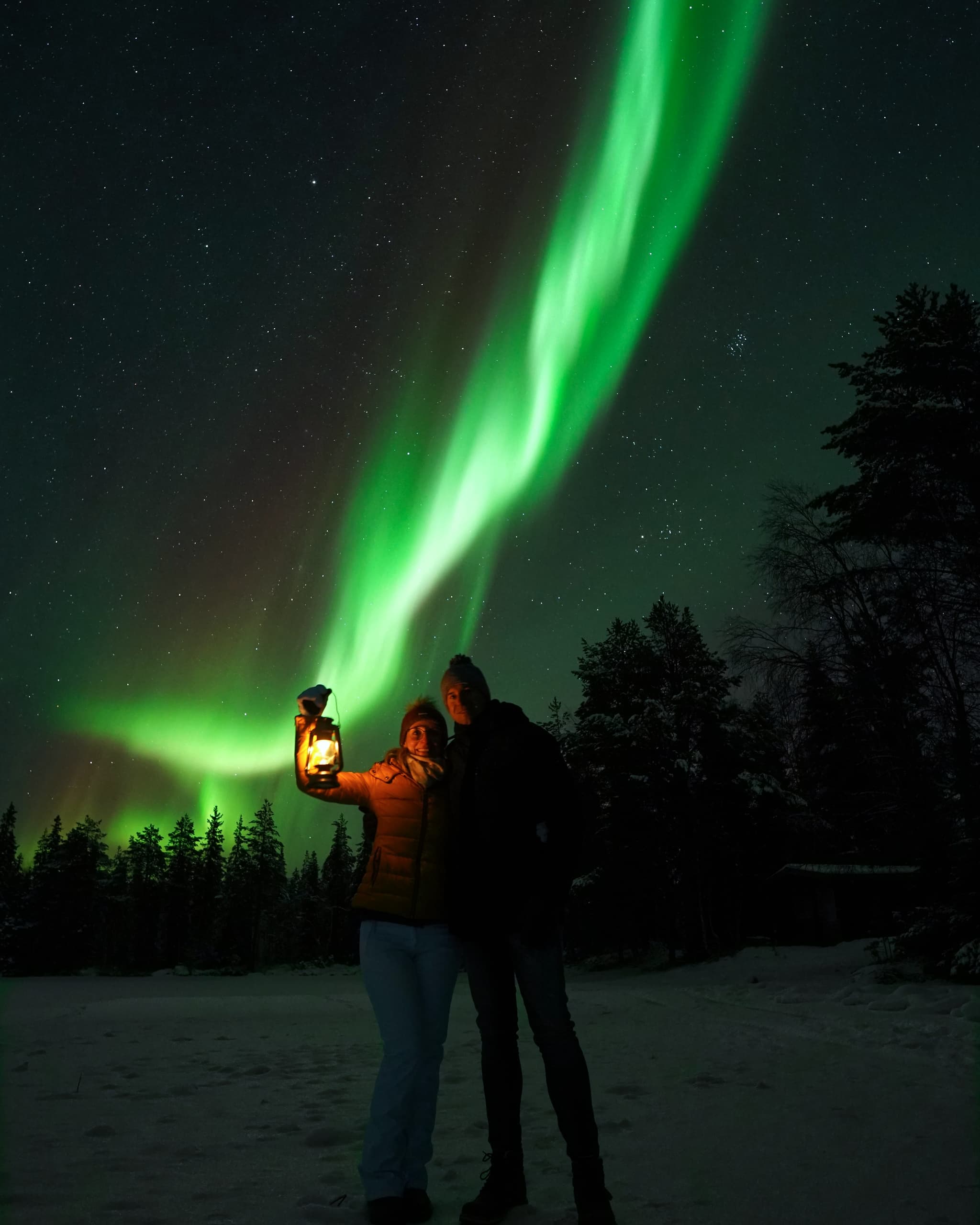 Two people stand in a snowy forest at night, one holding a lantern, beneath bright green northern lights and a starry sky.