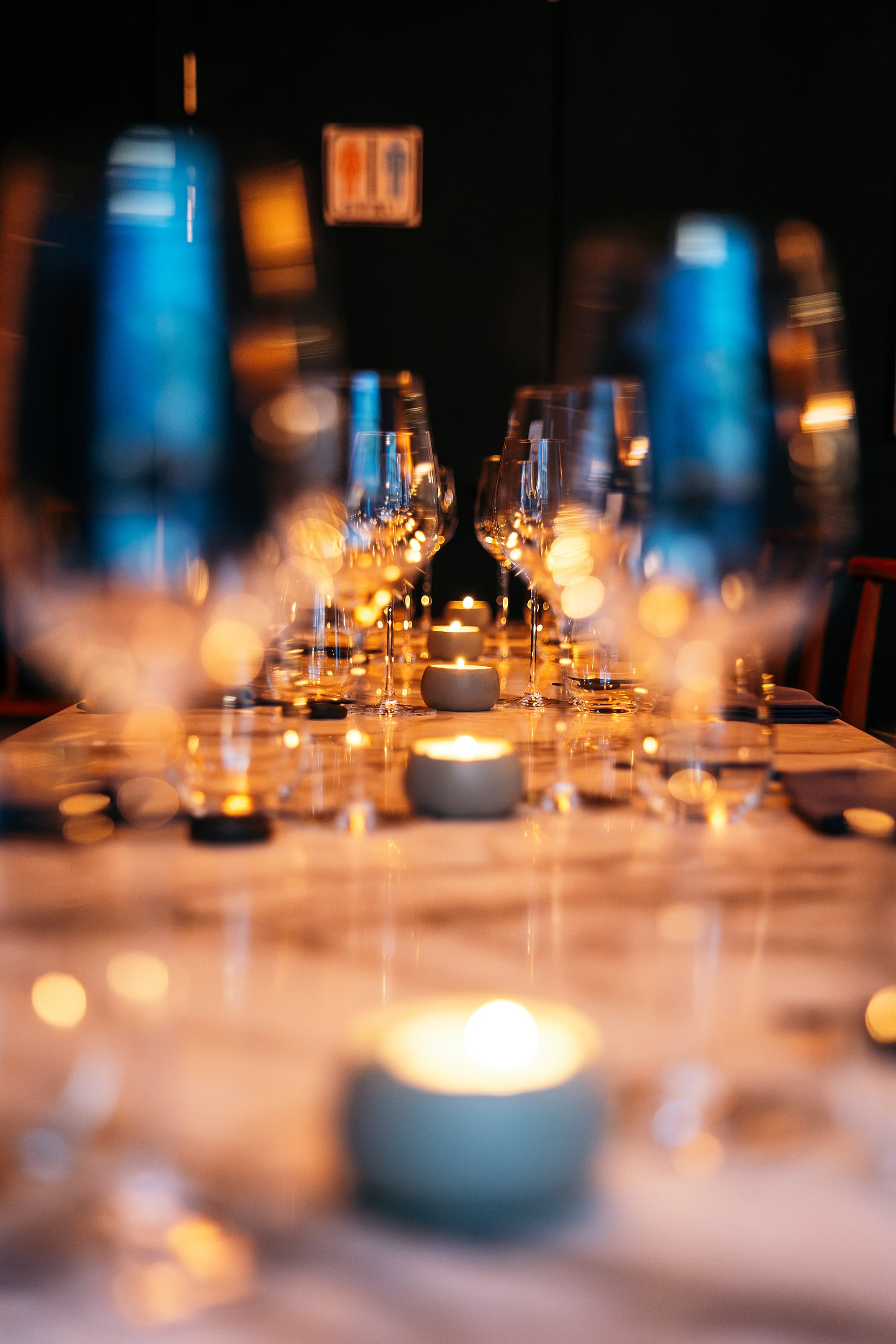 A candlelit dining table is set with rows of wine glasses and softly glowing votive candles in a dark interior.