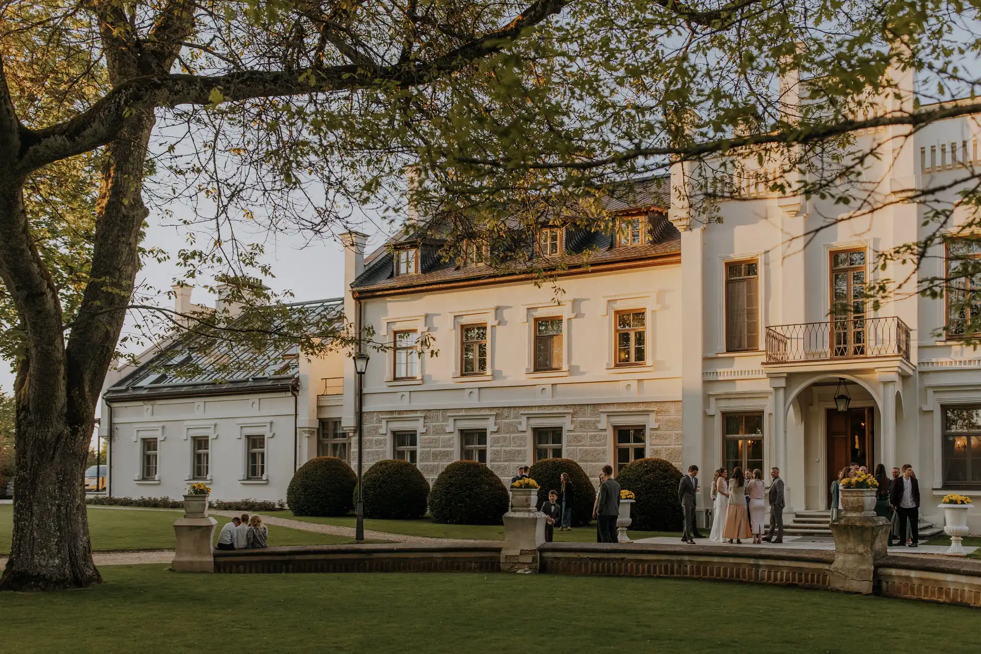 A stately manor-style building with manicured lawns and guests gathered outside under leafy trees in warm evening light.