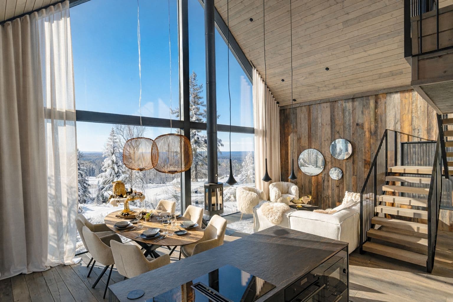 A bright modern chalet interior features a dining table, lounge seating, and a staircase beside floor-to-ceiling windows overlooking a snowy landscape.