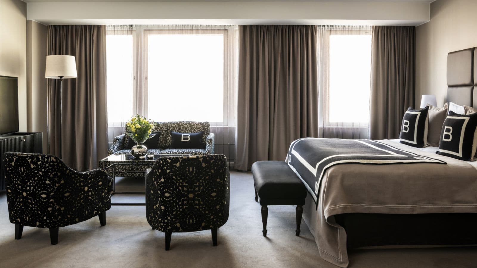 A stylish hotel suite features a king bed, patterned armchairs, a loveseat, and large windows with heavy curtains.