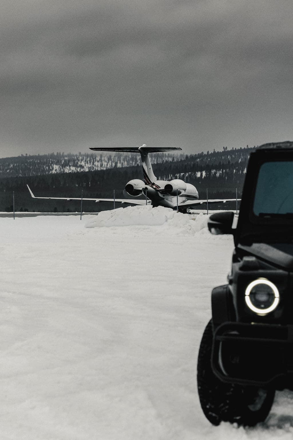 A private jet sits on a snowy airfield beside the front corner of a dark off-road vehicle under a gray winter sky.