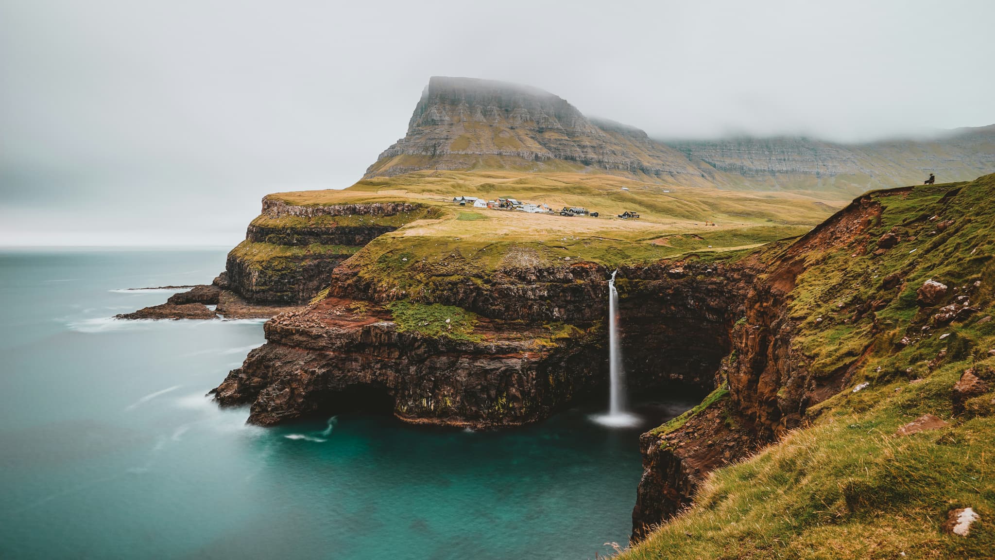 Coastal cliff landscape with a tall waterfall dropping into the sea, green grass slopes, and a misty mountain with a small settlement at its base.