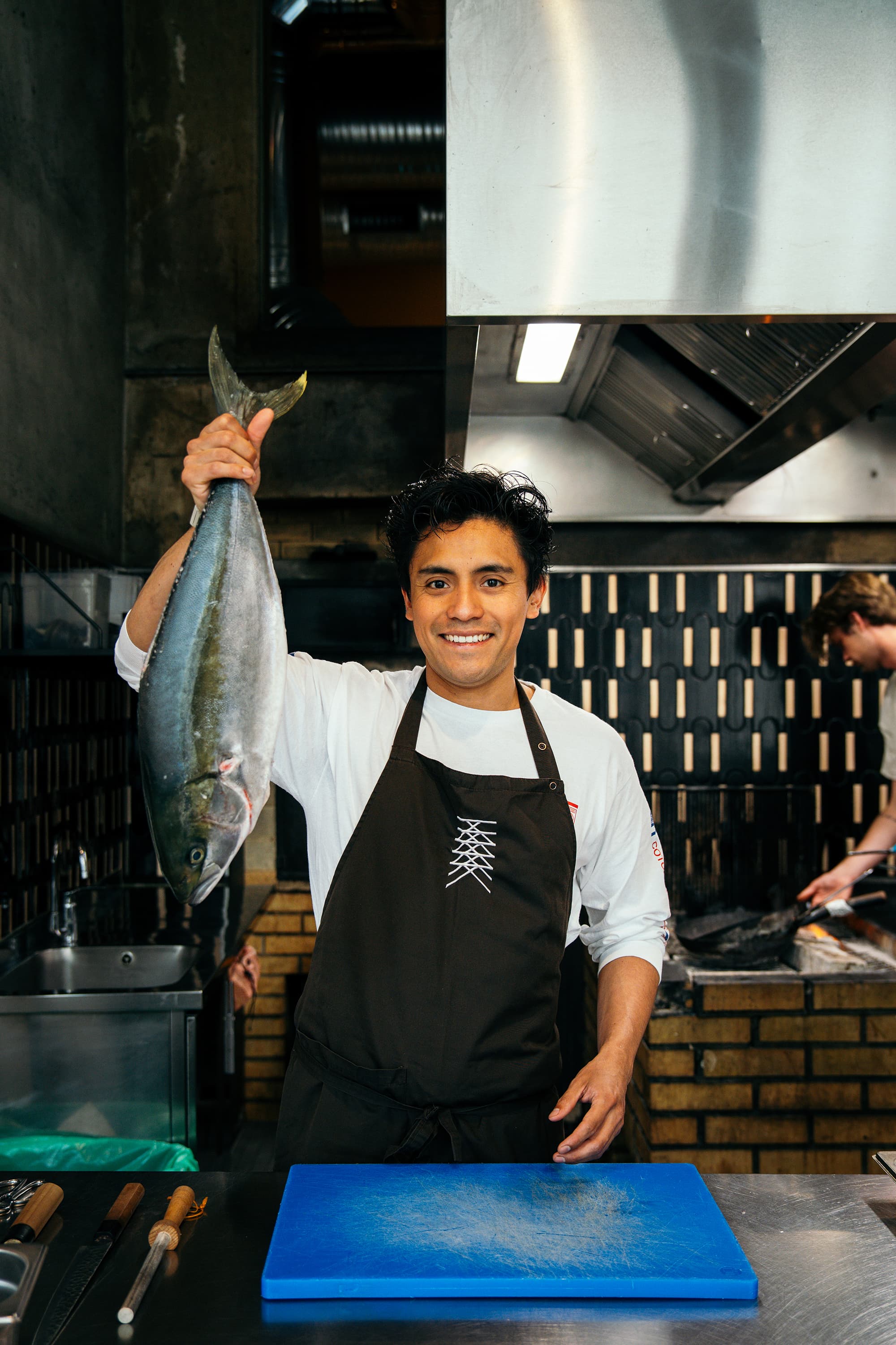 A smiling chef in a dark apron holds up a large whole fish in a commercial kitchen beside a blue cutting board.