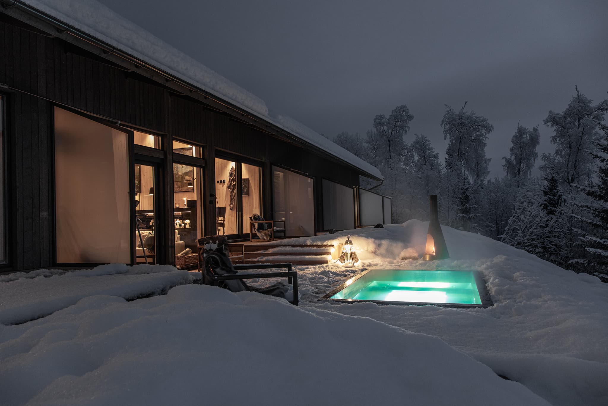 A modern wooden cabin with warm interior lighting sits in deep snow beside a glowing outdoor pool at dusk, surrounded by frosted trees.